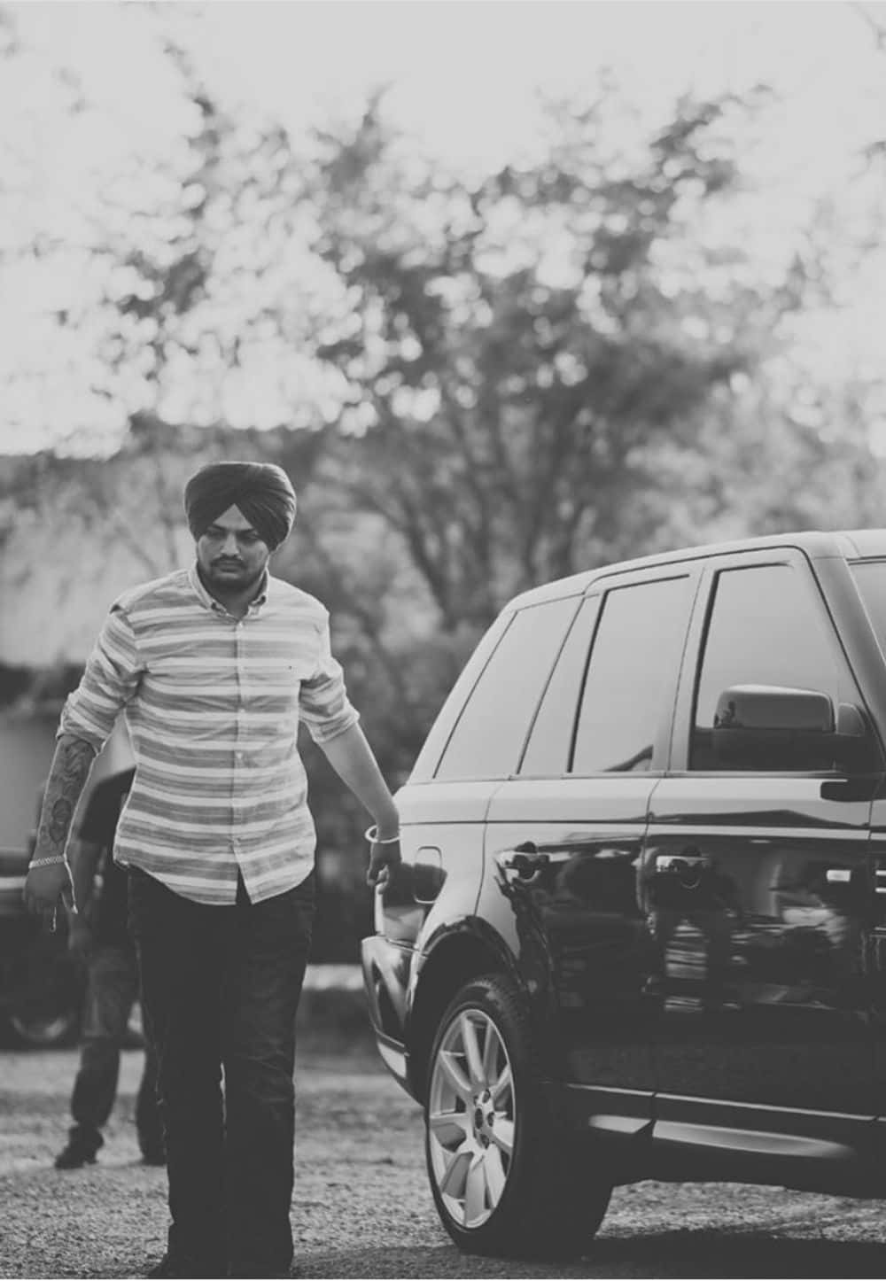 Sidhu Moose Wala 桌布 壁紙 — 一名戴頭巾的男子在柔和自然光下，背景有模糊的樹木，走在一輛時尚SUV旁邊。