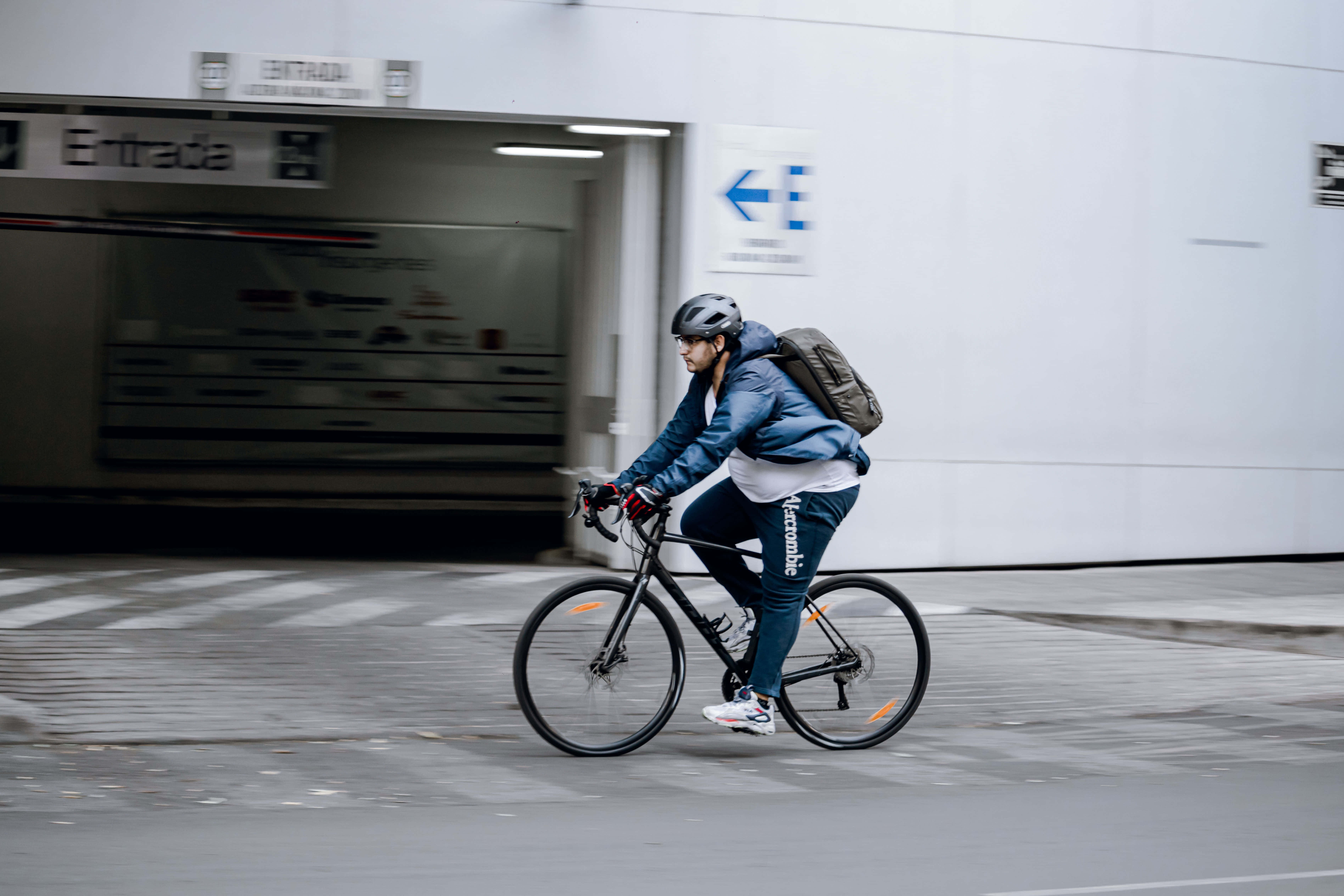Navy Blue Jacket 桌布 壁紙 — 一名戴頭盔和背囊的單車手在都市行人路上踩單車經過地鐵入口，帶有動態模糊效果。