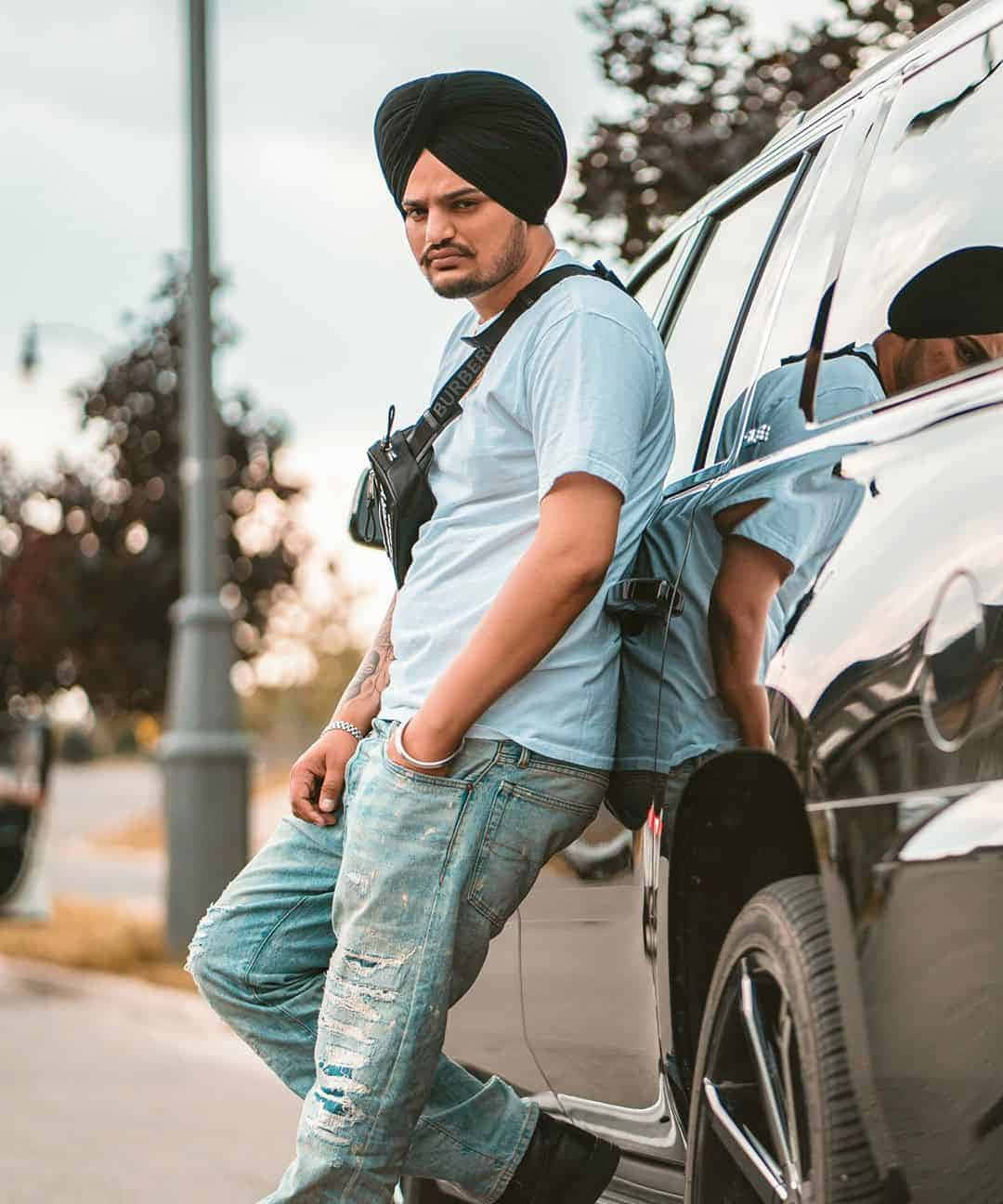 Sidhu Moose Wala 桌布 壁紙 — 一名戴黑色頭巾、穿破洞牛仔褲的男子，在種有樹木、光線柔和的街道上，靠在一輛閃亮的黑色SUV旁。
