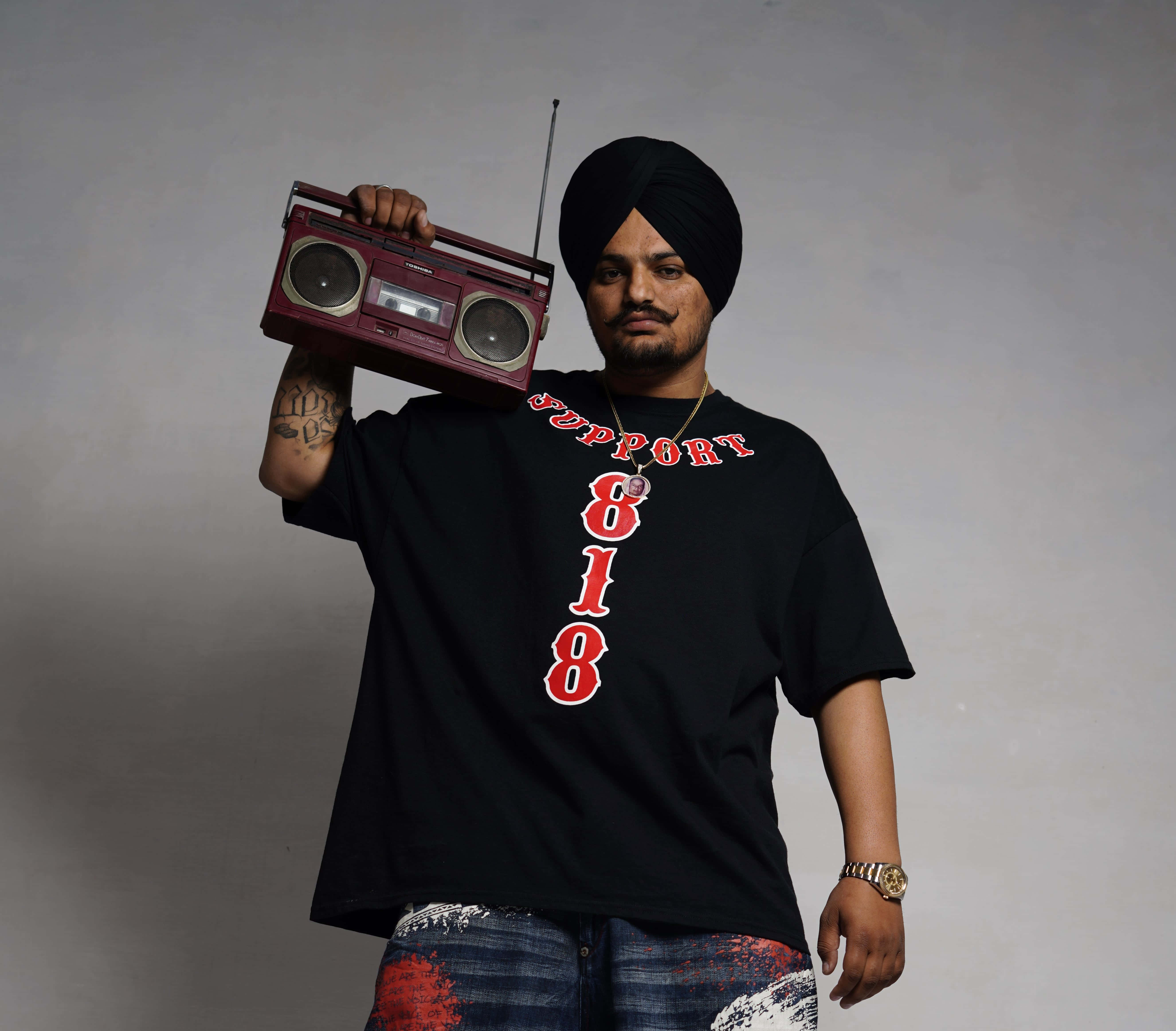 Sidhu Moose Wala 桌布 壁紙 — 一名戴黑色頭巾、身穿圖案T恤的男子，在純灰色攝影棚背景前拿著一個復古栗色手提音響。