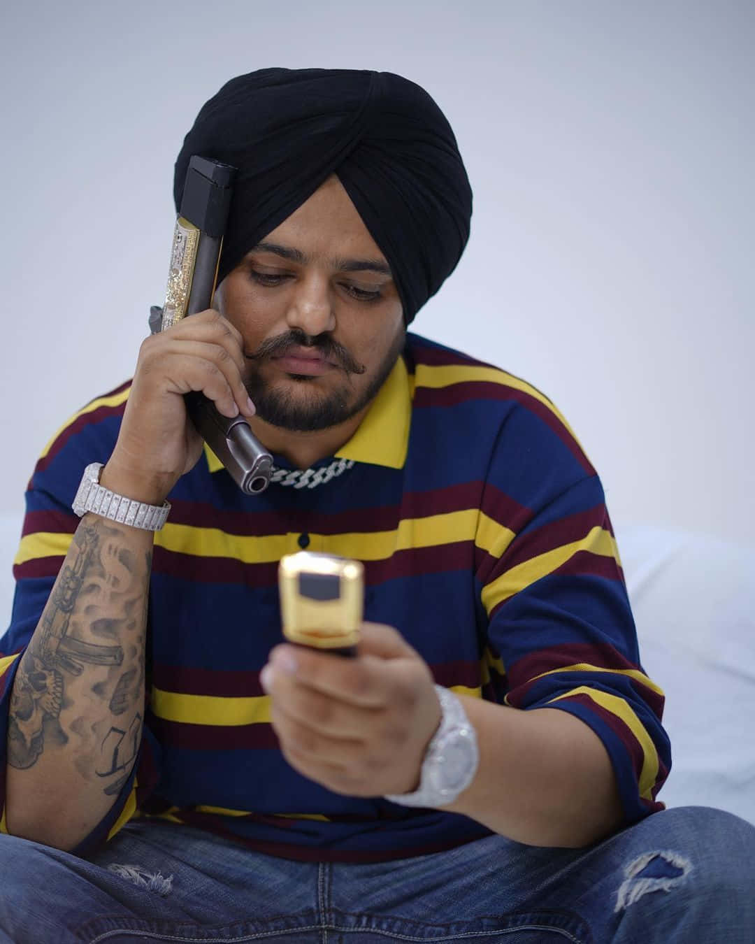 Sidhu Moose Wala 桌布 壁紙 — 一名戴黑色頭巾、身穿條紋襯衫的男子，將一部鍍金手機貼在耳邊，同時看著一部金色翻蓋手機。