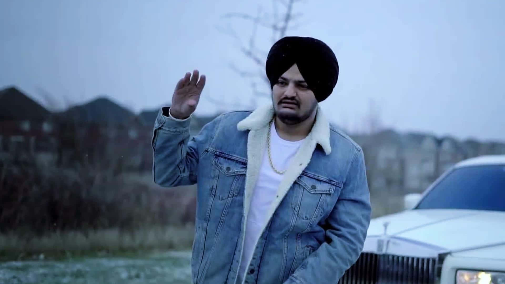 Sidhu Moose Wala 桌布 壁紙 — 一名戴黑色頭巾、身穿牛仔褸的男子，在荒涼的郊區景觀中，於一輛白色豪華房車旁揮手。