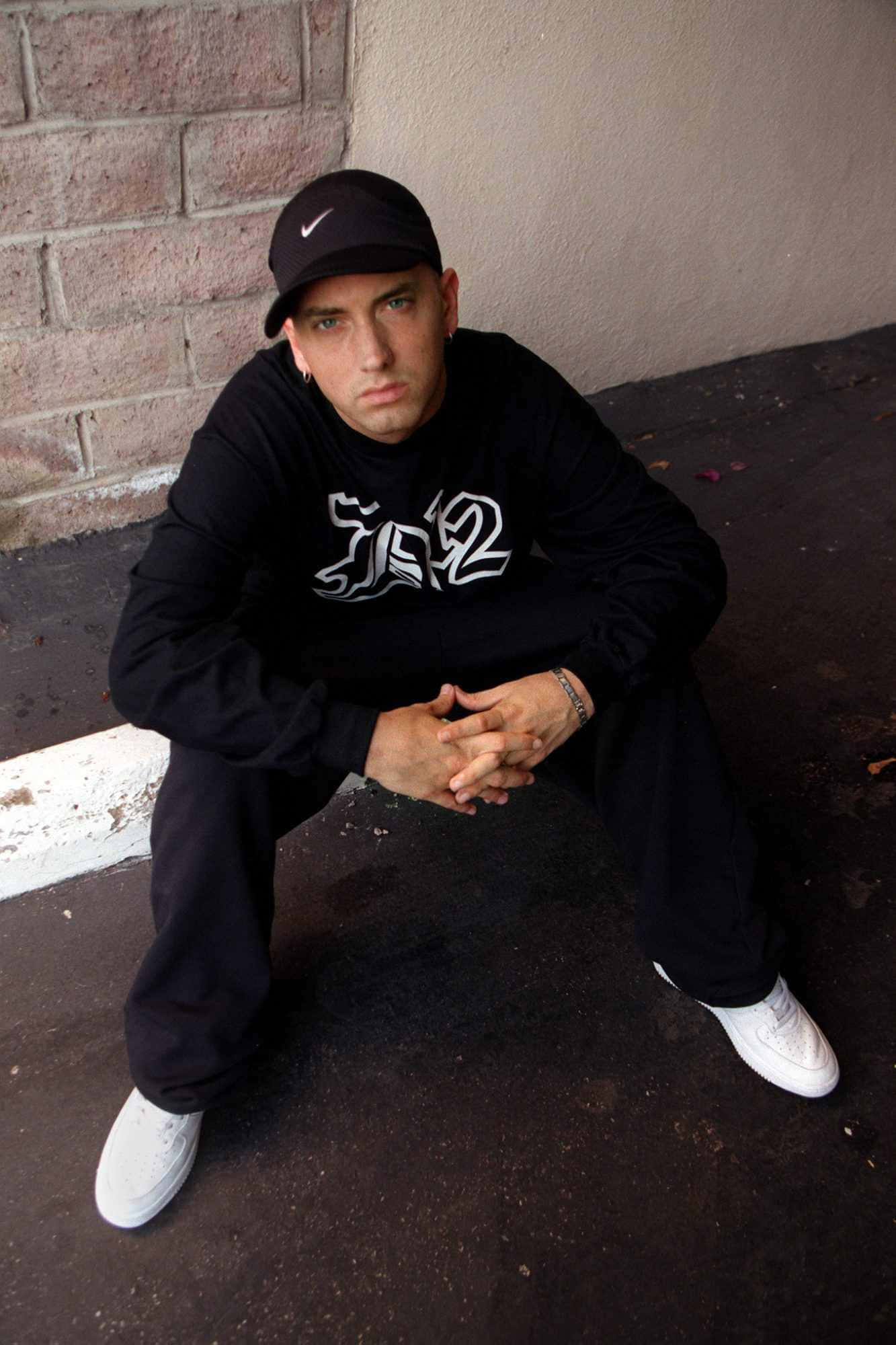Eminem 桌布 壁紙 — 一名戴黑色鴨舌帽、穿黑色運動衫的年輕男子蹲在磚牆旁，眼神凌厲。