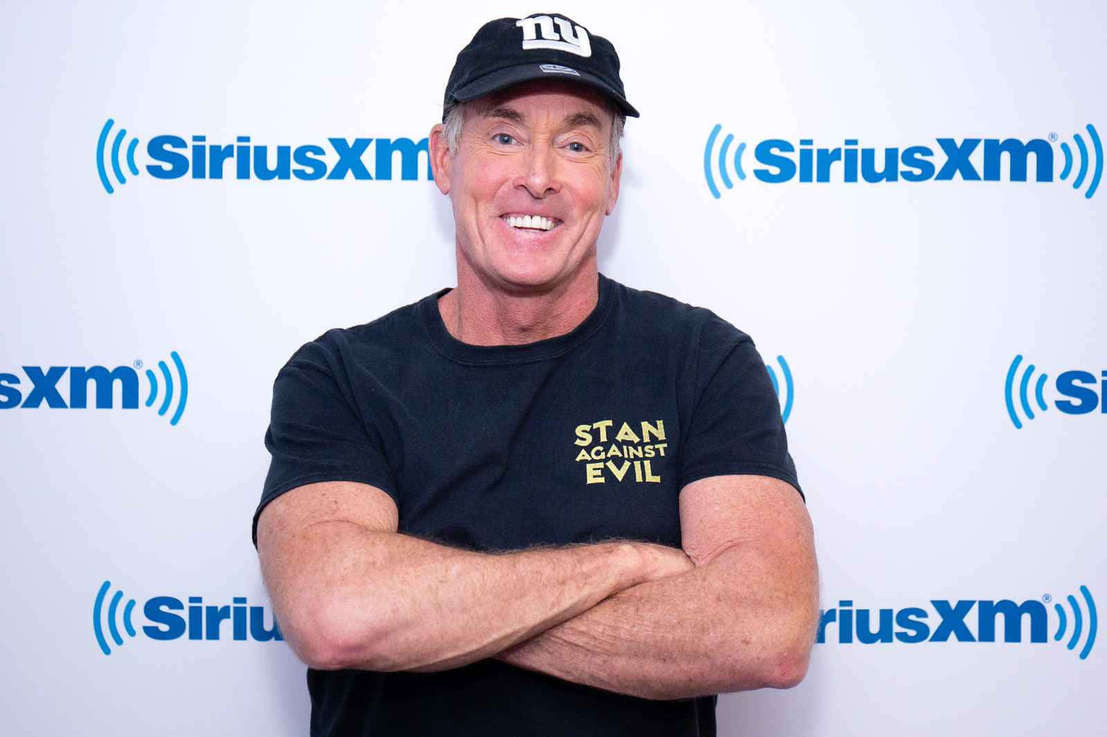 John C. Mcginley 桌布 壁紙 — 一名戴黑色鴨舌帽和T恤的男子微笑著，自信地交叉雙臂站在SiriusXM品牌背景前。