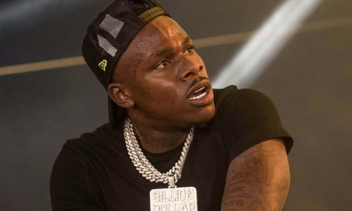 Dababy 桌布 壁紙 — 一名戴黑色鴨舌帽和鏈條項鍊的男子在明亮的舞台燈光下向上看，表情強烈。