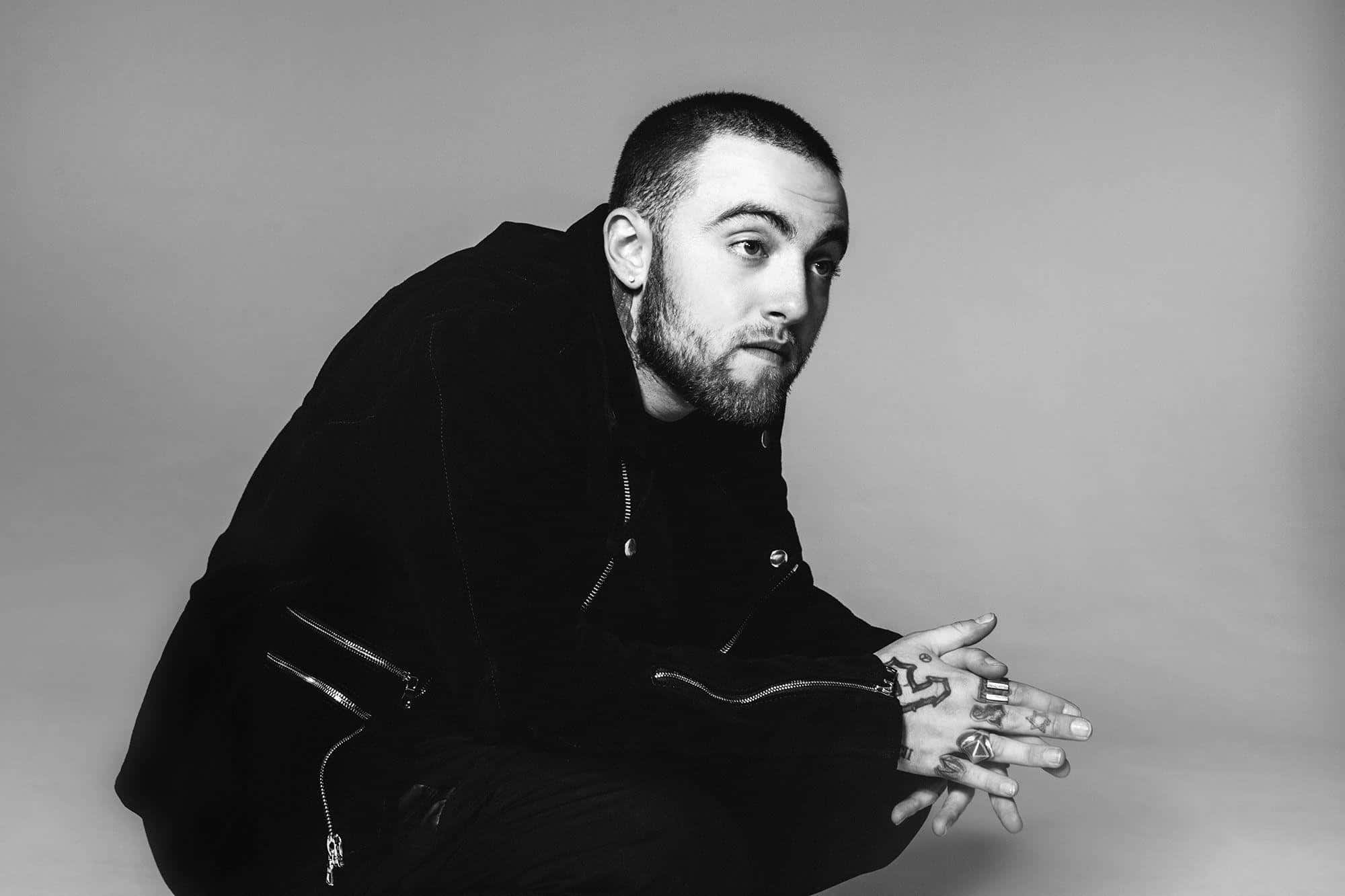 Mac Miller 桌布 壁紙 — 一名手上有紋身的男子穿著黑色衣服，在單色調的純灰色背景前蹲下。