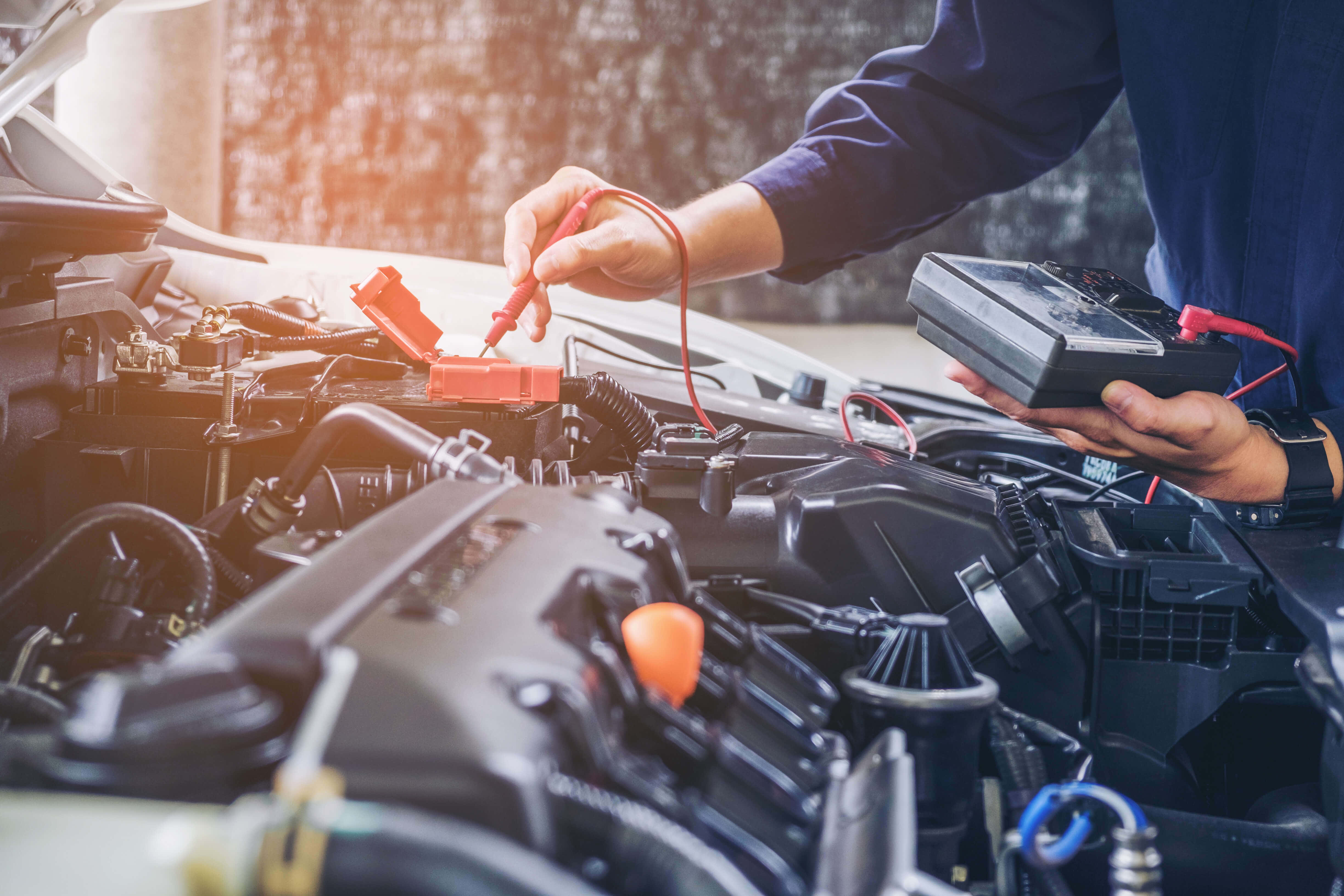 Car Repair 桌布 壁紙 — 一名技師在打開的引擎蓋附近，於溫暖陽光下用萬用錶測試汽車電池。