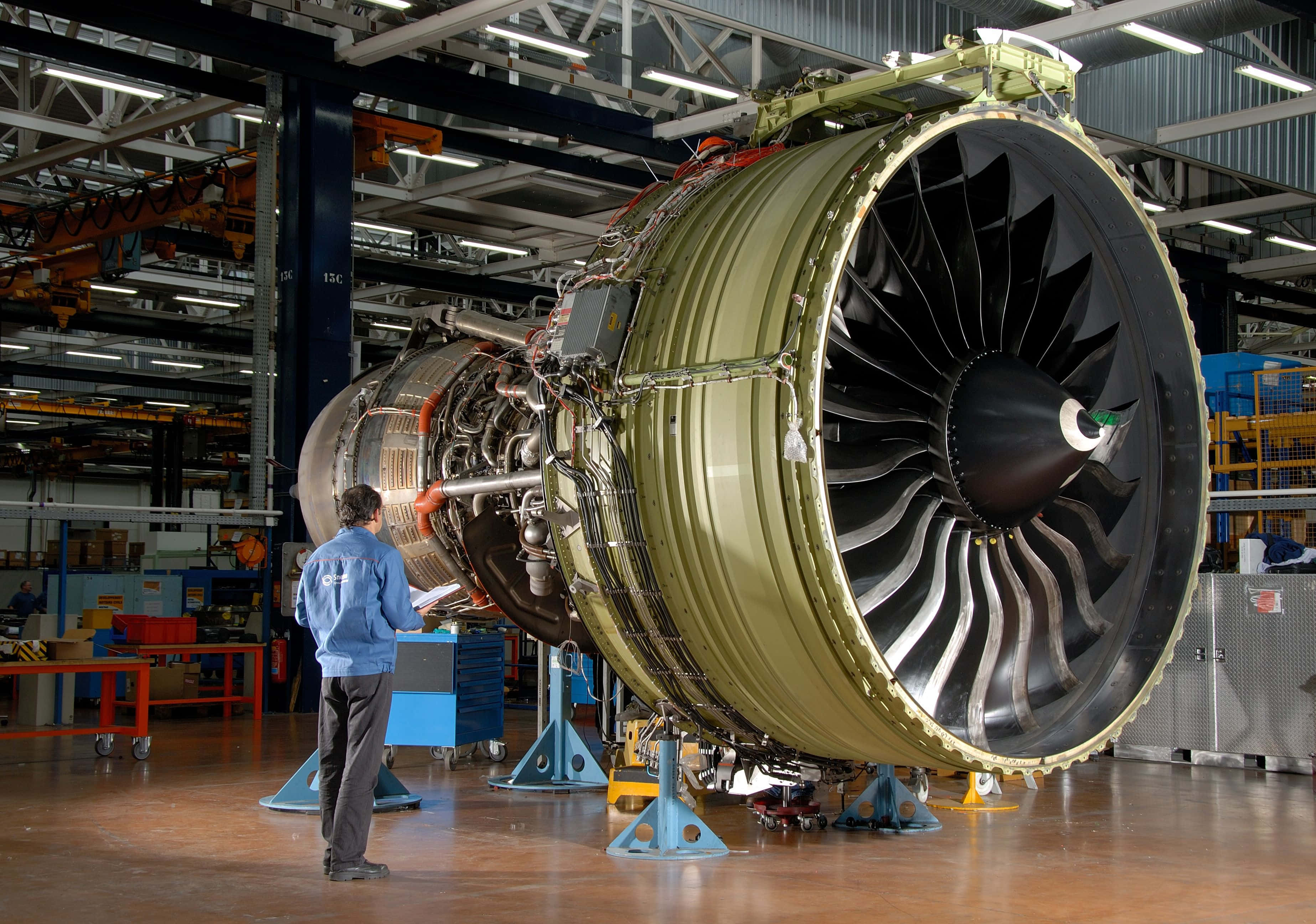 Jet Engine 桌布 壁紙 — 一名技術人員在光線充足的工業機庫內檢查巨大的噴射引擎，機庫內有高架起重機和工具車。