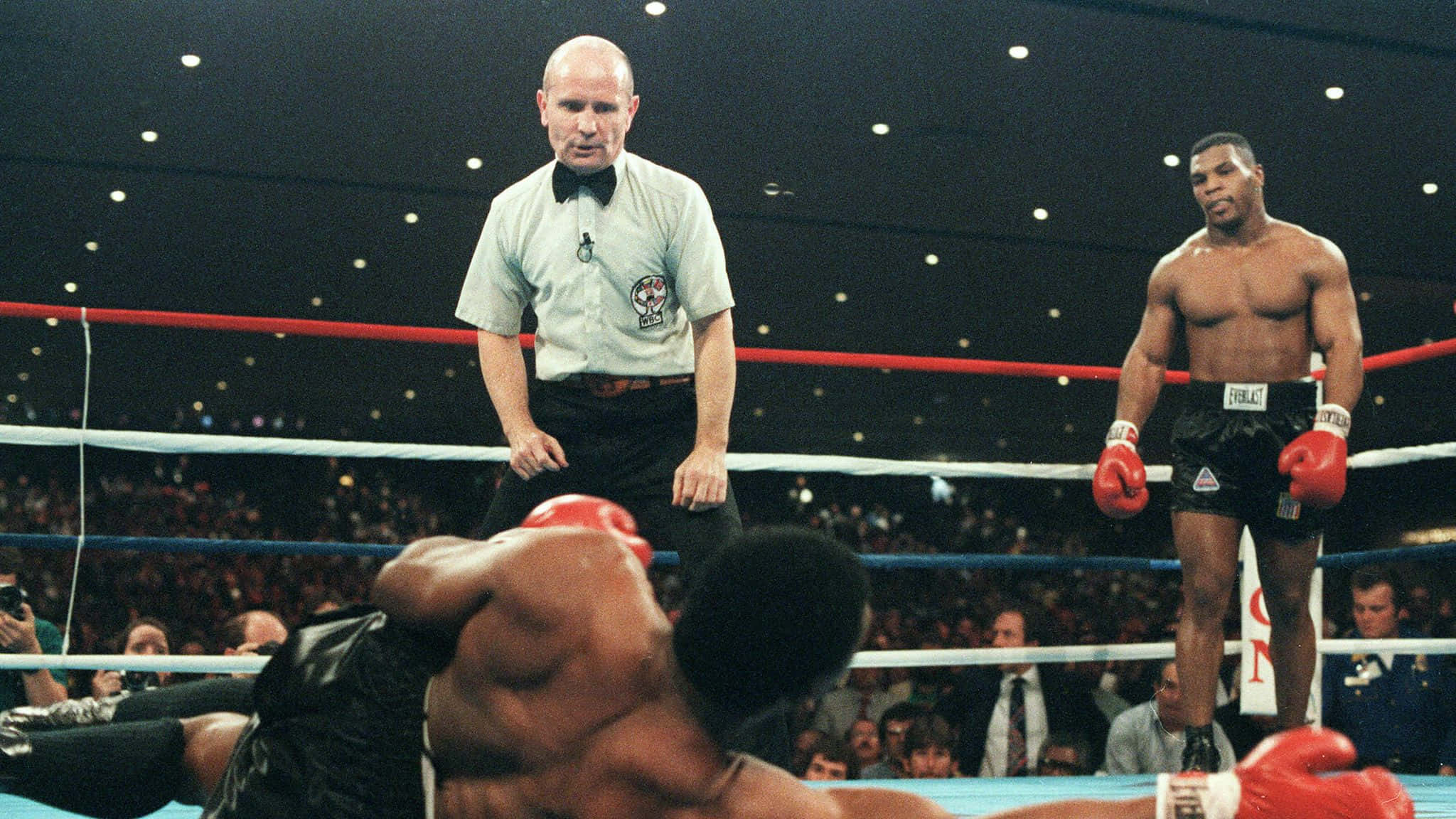 mike tyson 桌布 壁紙 — 一名拳擊手在擂台上被擊倒在帆布上，對手勝利地站著，裁判專注地觀看。