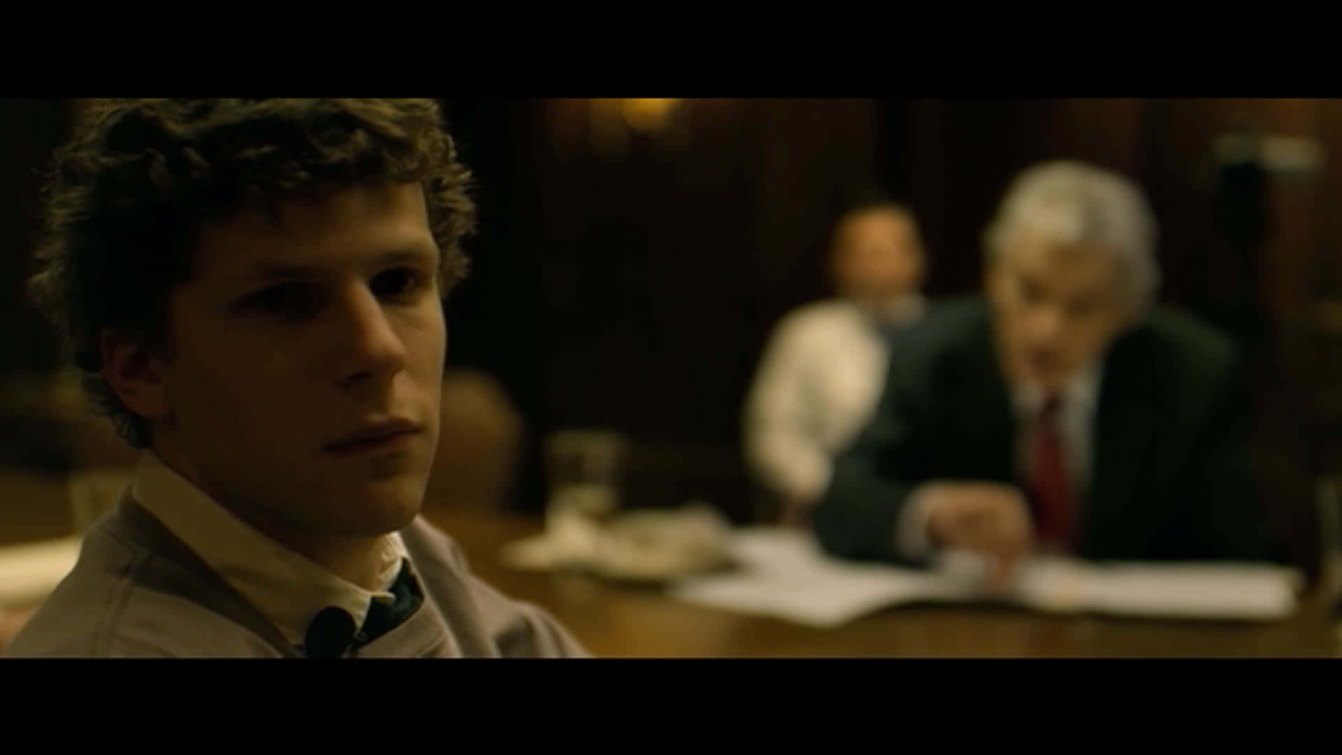 the social network 桌布 壁紙 — 一名捲髮年輕男子在燈光昏暗的法庭內專注地凝視，背景中一名身穿西裝的年長男子坐在木桌旁。