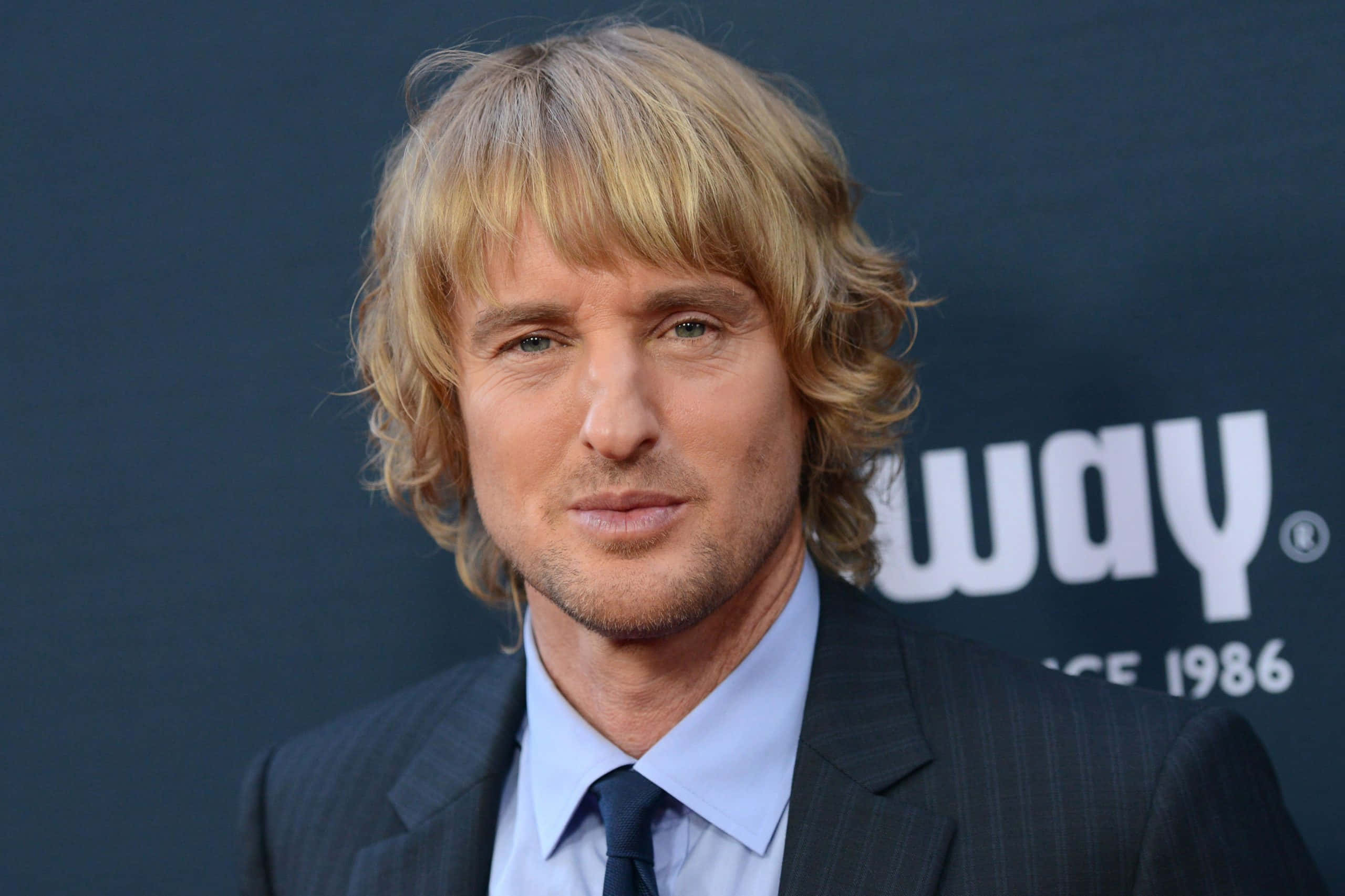 Owen Wilson 桌布 壁紙 — 一名擁有波浪形金髮和藍眼睛的男子，身穿深色細條紋西裝和領帶，背景為深色並帶有白色文字。