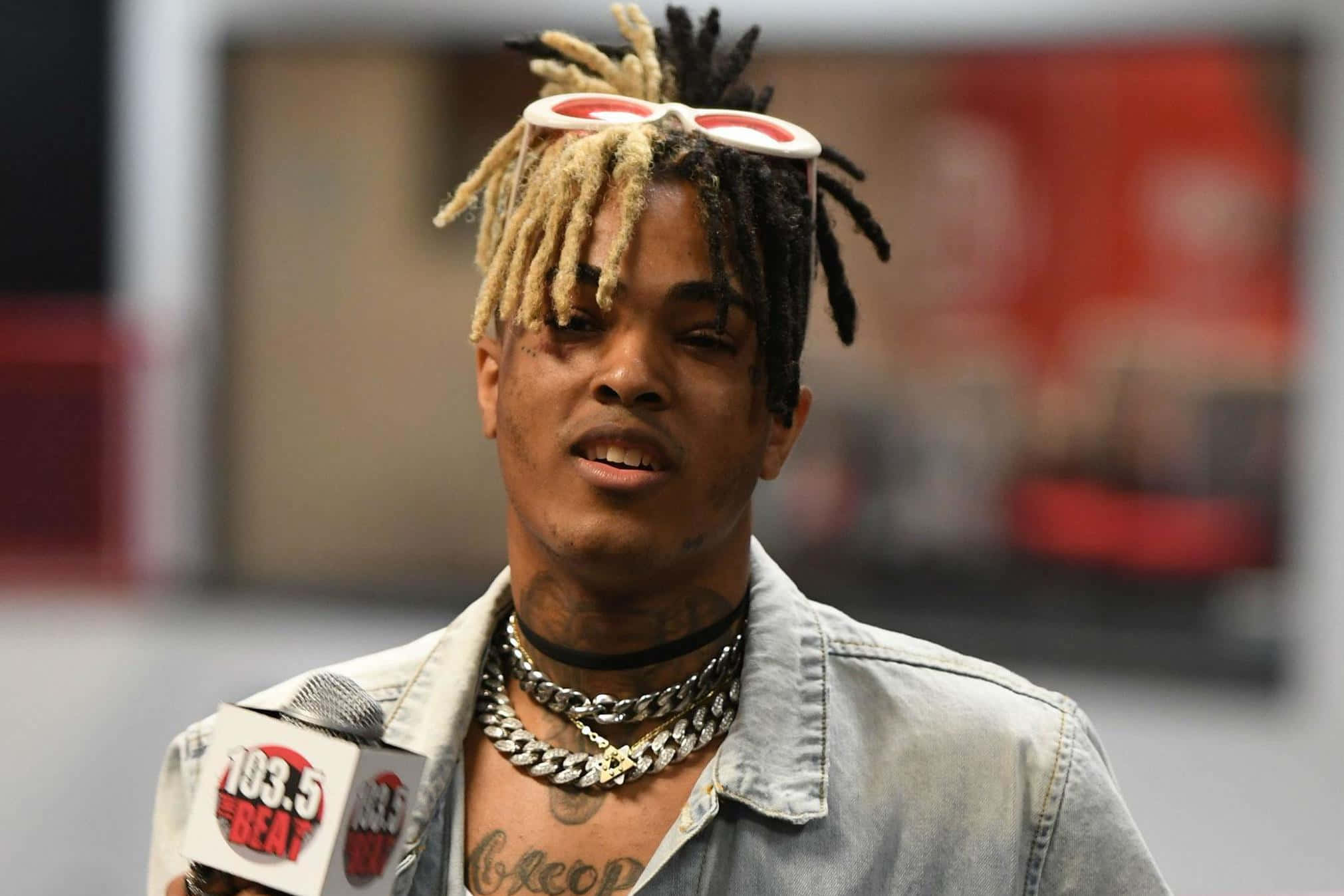 XXXTENTACION 桌布 壁紙 — 一名擁有金色和黑色辮子頭的男子，頭上戴著白色鏡框眼鏡，佩戴多層銀鏈，正在對著麥克風說話的特寫。