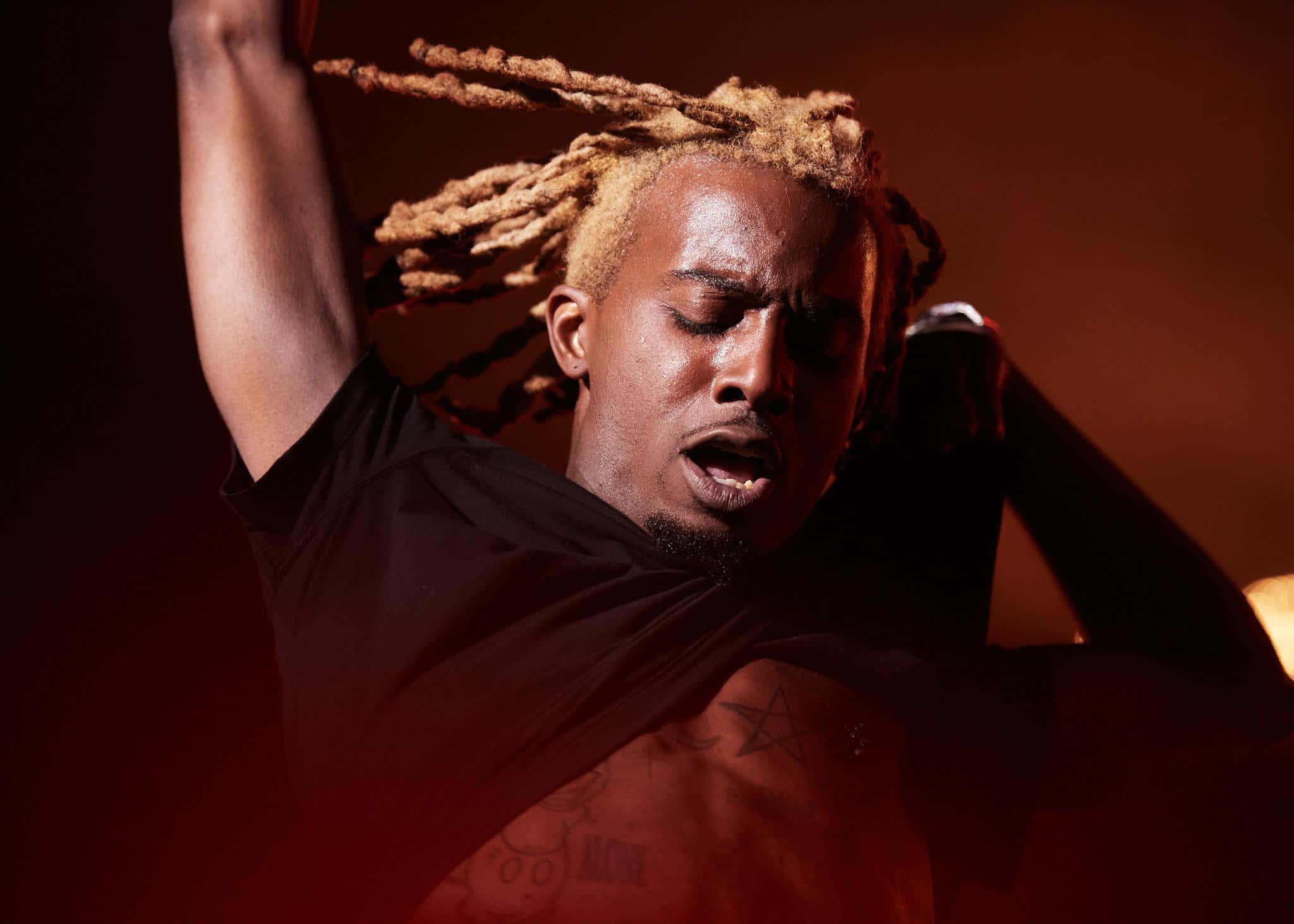 playboi carti 桌布 壁紙 — 一名擁有金色辮子辮的男人在溫暖的舞台燈光下熱情歌唱，胸前的紋身清晰可見。