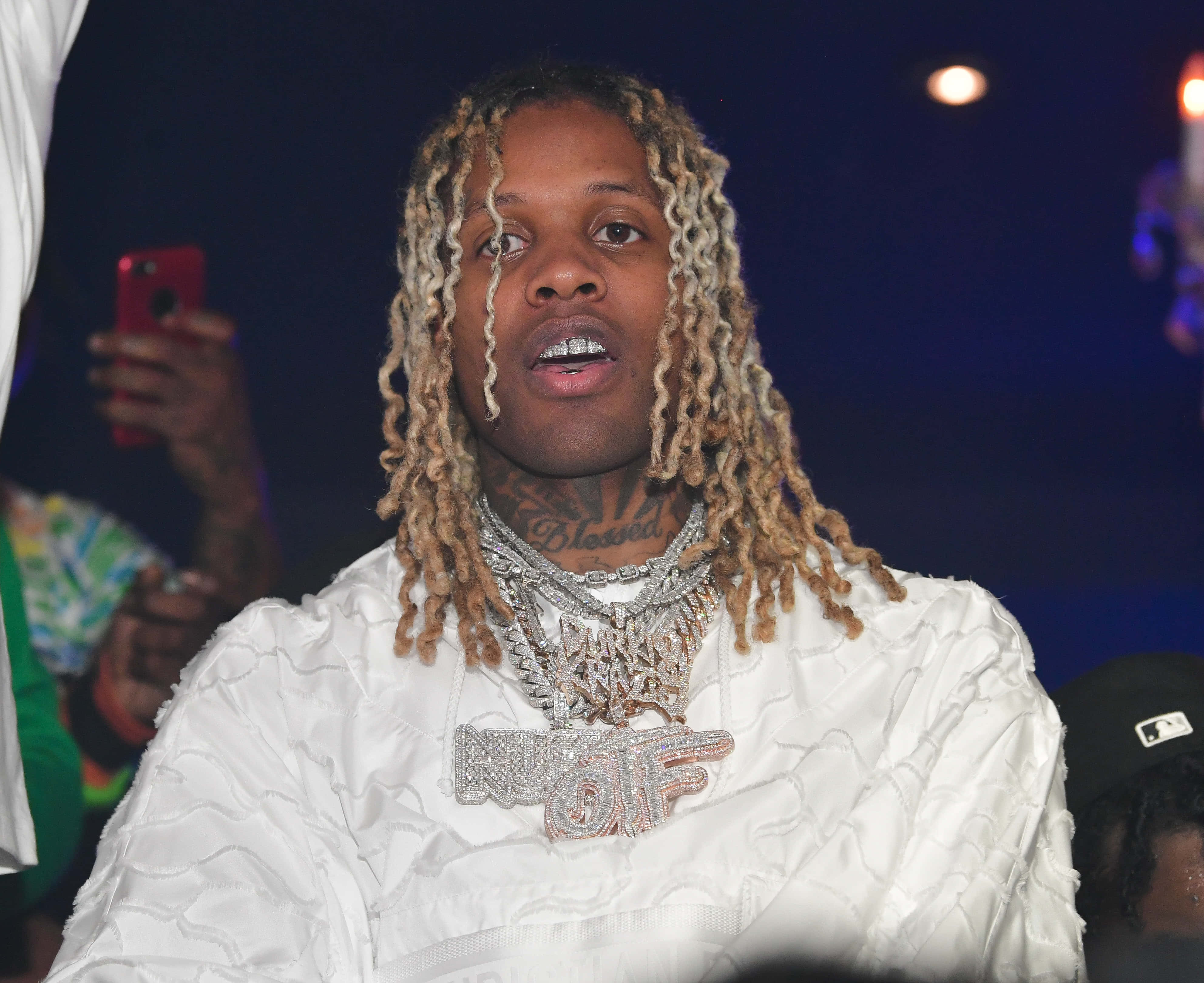 lil durk 桌布 壁紙 — 一名擁有金色辮子頭和沉重銀鏈的男子，在舞台燈光下穿著白色紋理外套。