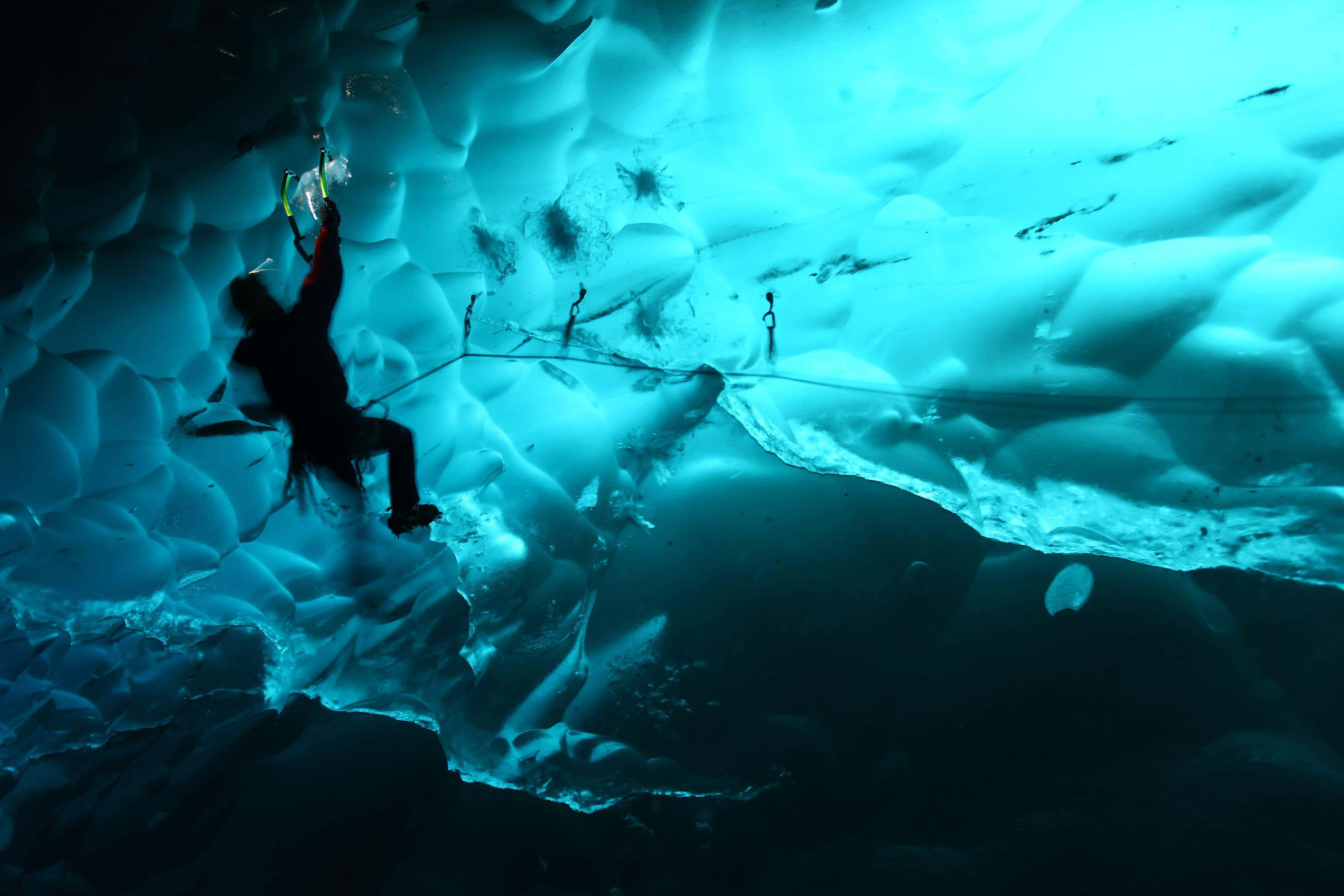 Ice Climbing 桌布 壁紙 — 一名攀岩者在戲劇性燈光下，使用繩索裝備攀登發光藍色冰洞天花板。