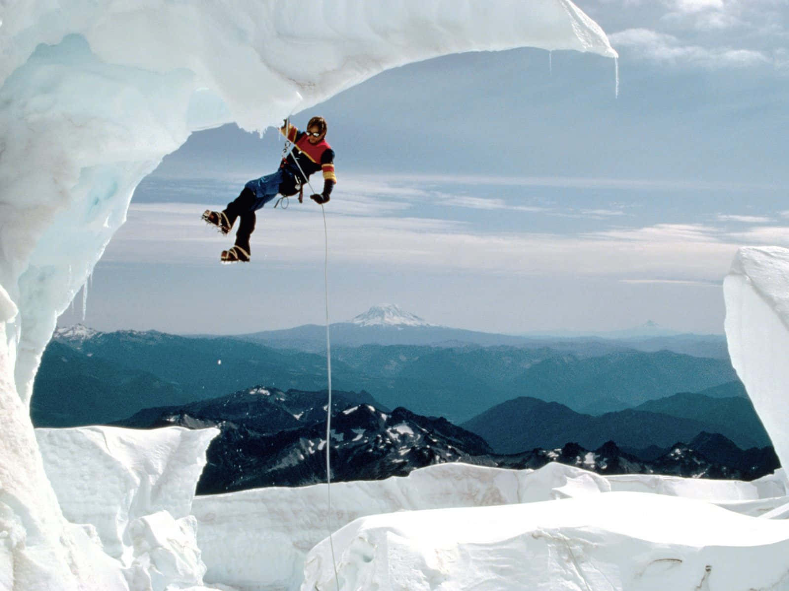 Ice Climbing 桌布 壁紙 — 一名攀岩者攀爬一個巨大的冰拱門，下方可見山脈和白雪覆蓋的山峰，天空清澈湛藍。