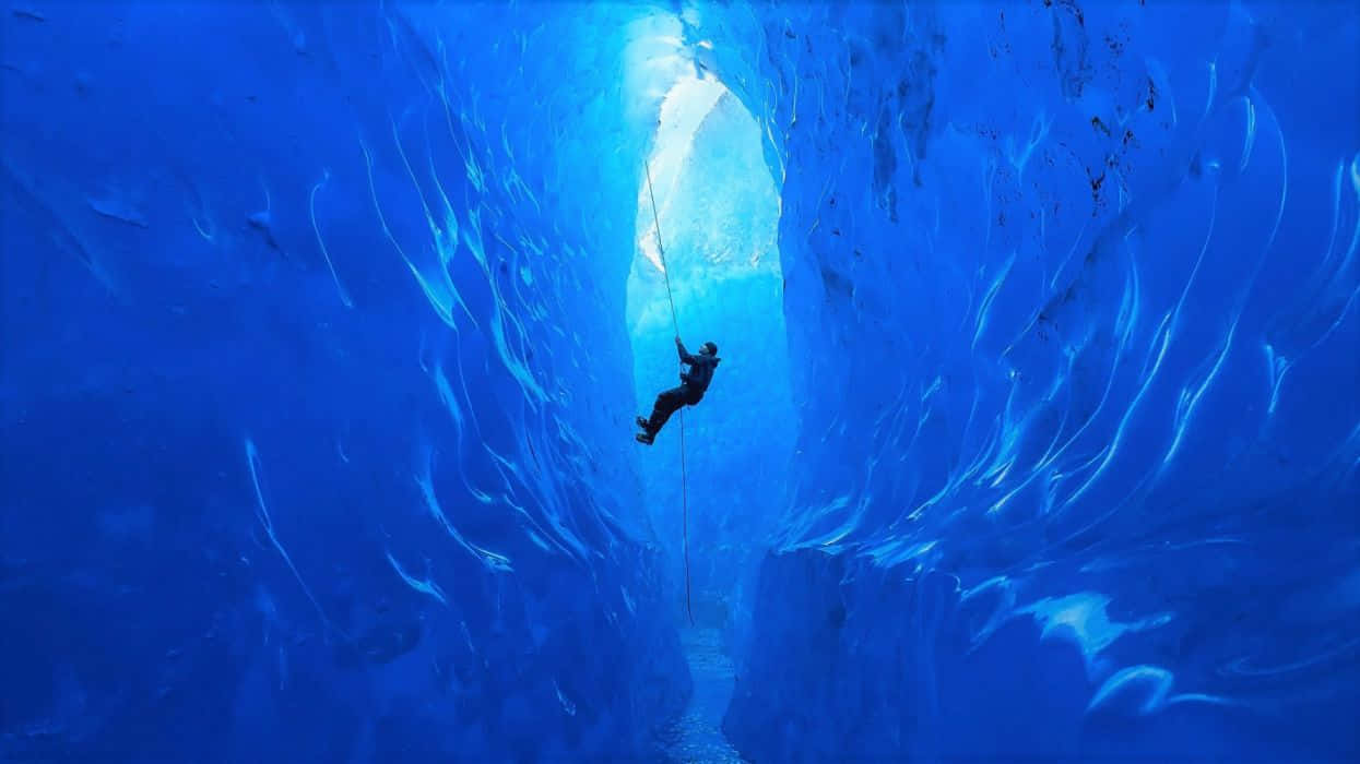 Ice Climbing 桌布 壁紙 — 一名攀岩者正沿著高聳的藍色冰洞下降，陽光從上方的冰層天花板傾瀉而下。