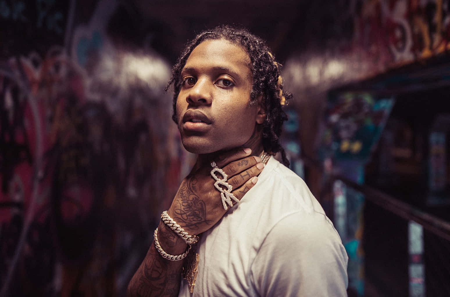 lil durk 桌布 壁紙 — 一名有紋身和戴金鏈的男子在昏暗的城市環境中，自信地以塗鴉牆為背景擺姿勢。