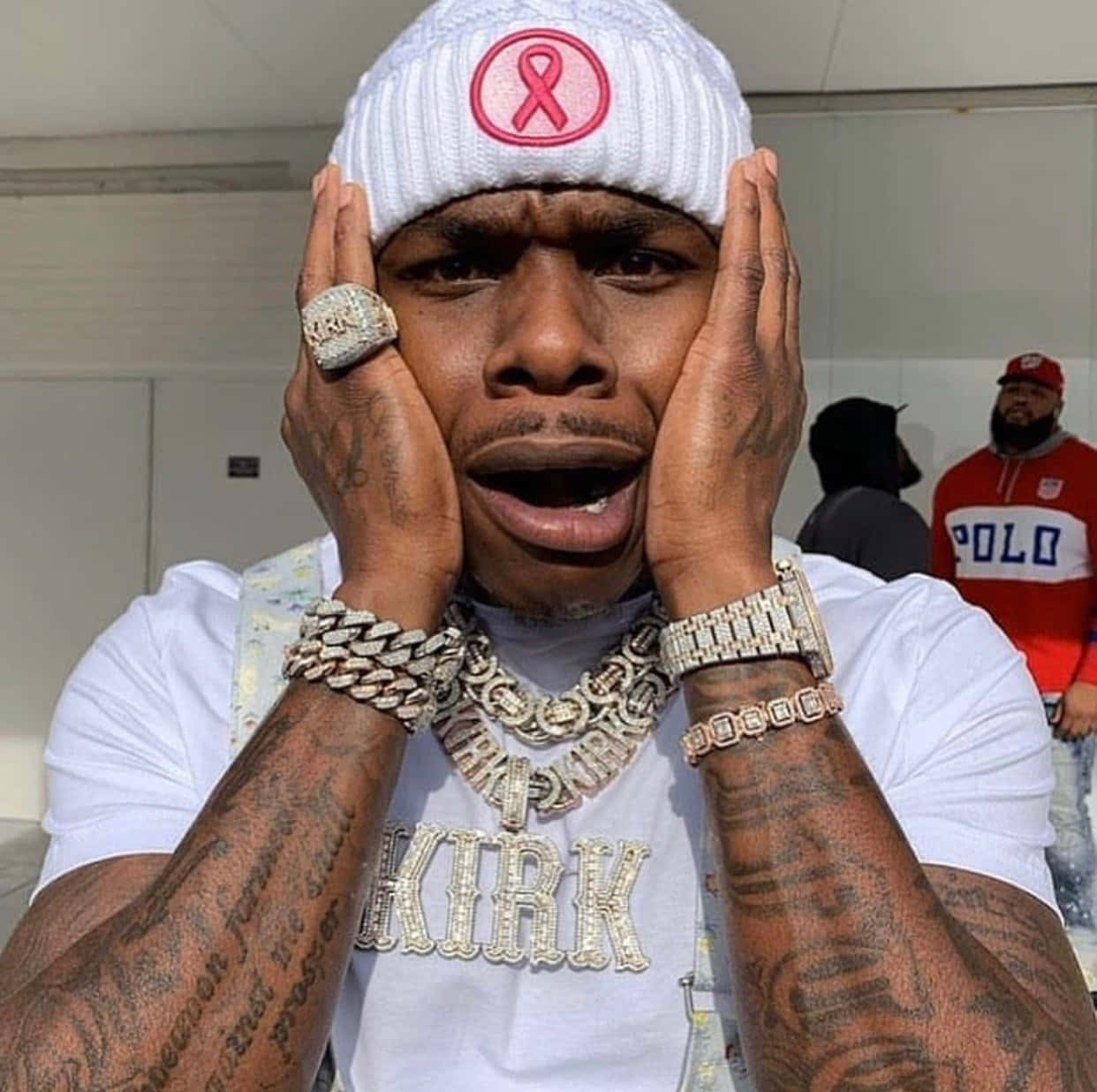 Dababy 桌布 壁紙 — 一名有紋身手臂的男子戴著白色無邊帽，帽上有粉紅色絲帶，佩戴鑲鑽珠寶，身穿印有「KIRK」的白色襯衫，震驚地捧著臉。