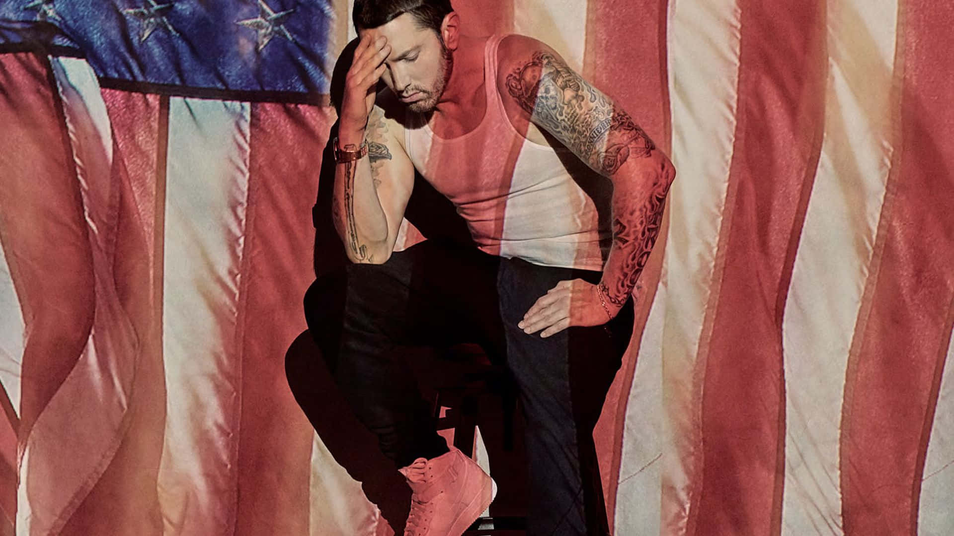 Eminem 桌布 壁紙 — 一名有紋身的男子身穿白色背心，跪在垂掛的美國國旗前，手放在額頭上，陷入沉思。