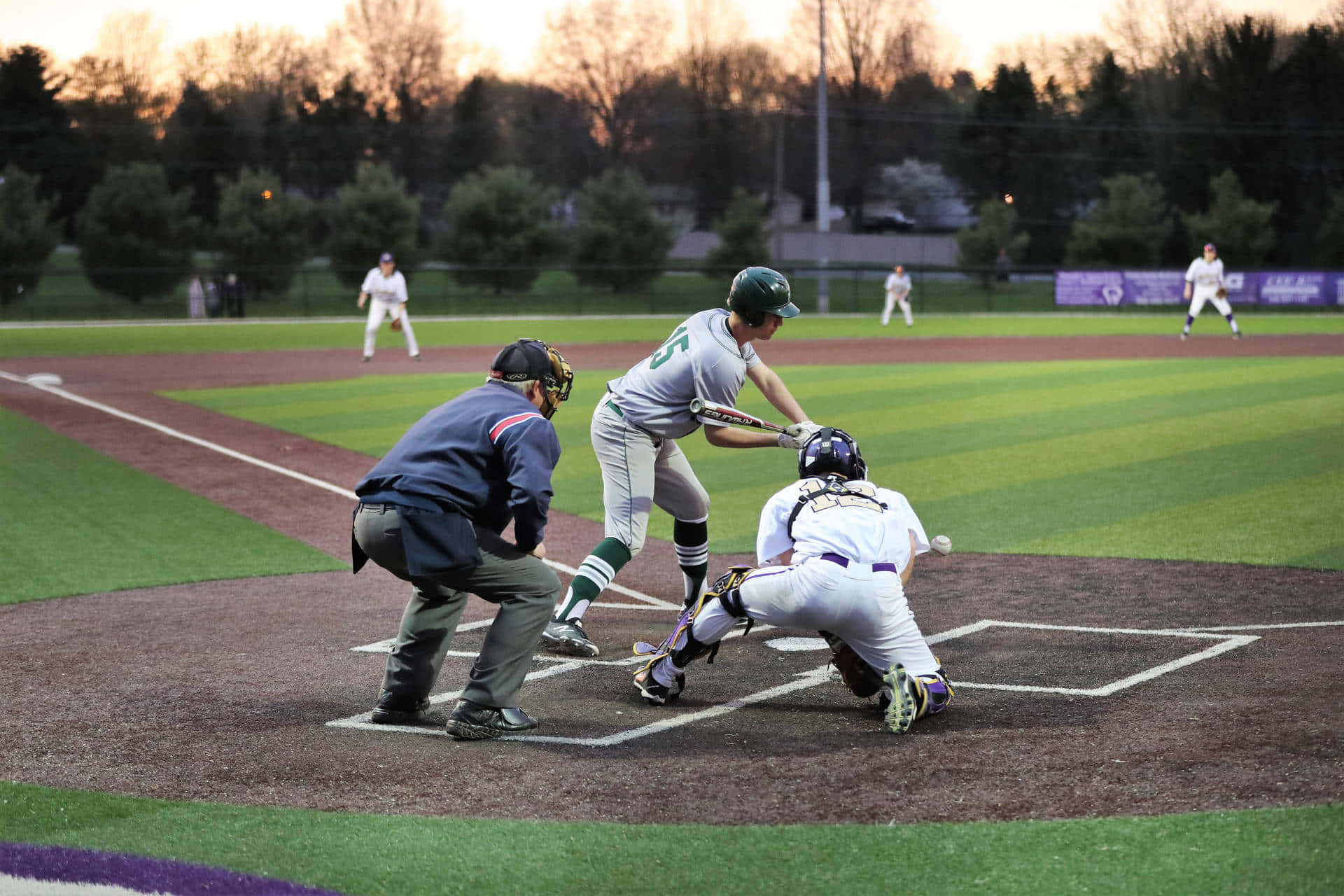 College Baseball 桌布 壁紙 — 一名棒球打擊手在黃昏時揮棒，捕手和裁判蹲在本壘板後方，場地維護良好。