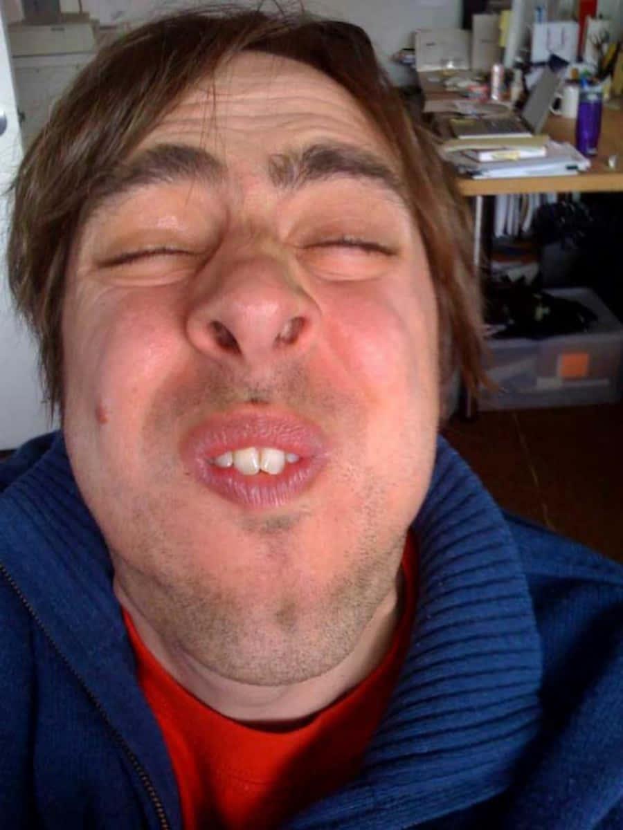 Funny Faces 桌布 壁紙 — 一名棕色頭髮、身穿紅色襯衫的男子在雜亂的房間裡閉著眼睛、噘起嘴唇，做出一張傻臉。