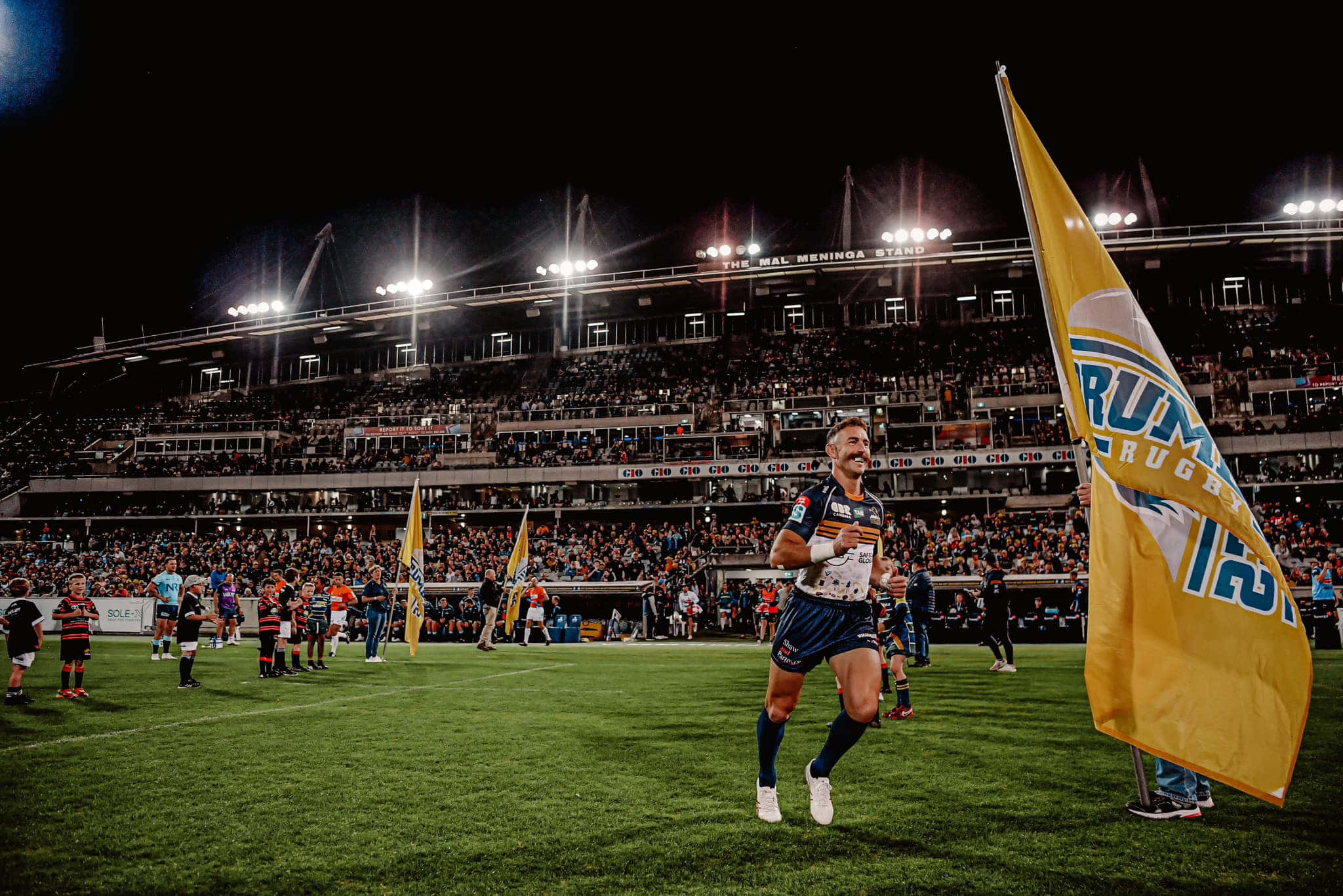 Brumbies 桌布 壁紙 — 一名欖球運動員在夜間球場燈光下慶祝勝利，看台上球迷揮舞旗幟。