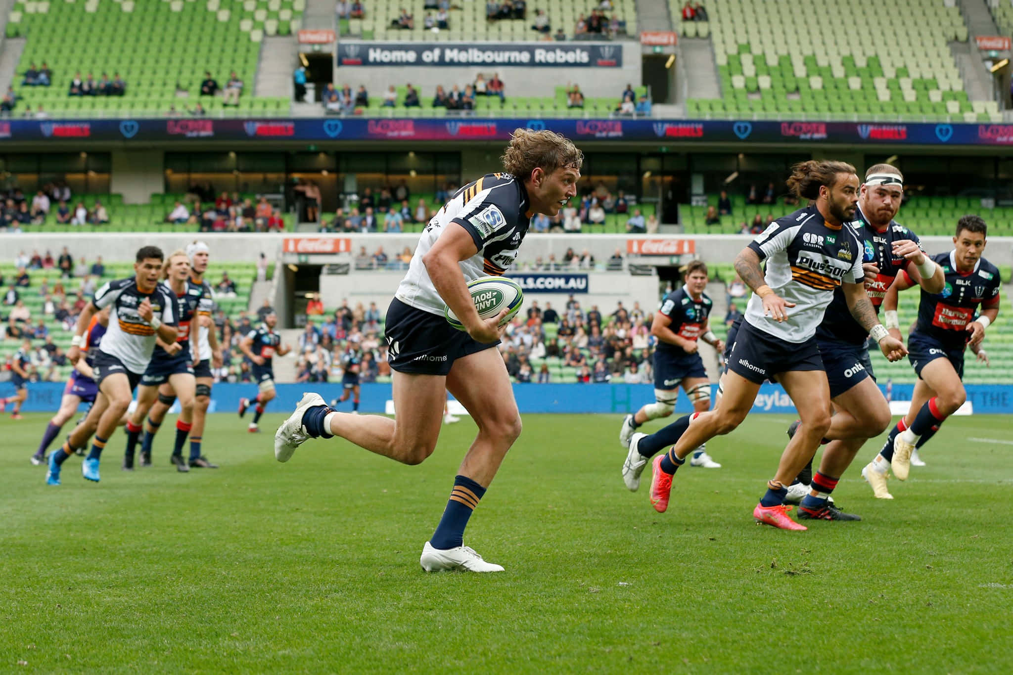 Brumbies 桌布 壁紙 — 一名欖球運動員帶球衝刺，隊友和對手在明亮的日光下於充滿活力的綠色球場上追趕他。