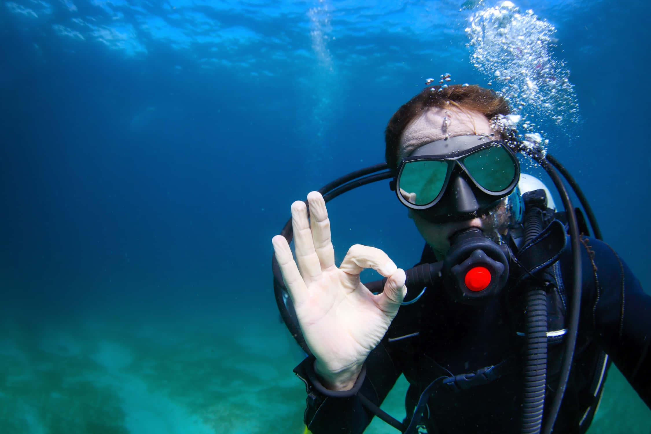 diving 桌布 壁紙 — 一名水肺潛水員在水下做出OK手勢，周圍有氣泡在清澈的藍色海水中升起。