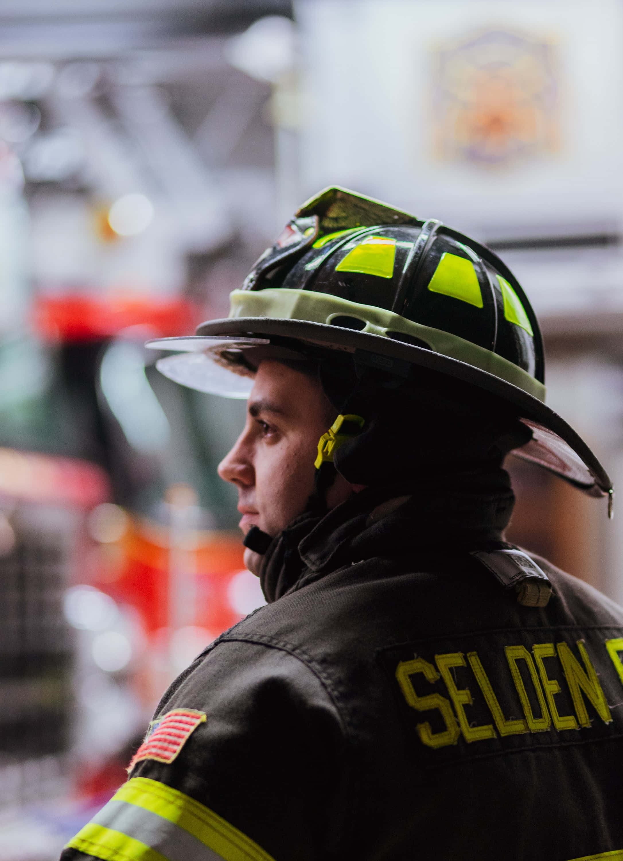 消防員 桌布 壁紙 — 一名消防員側面照，戴著頭盔和制服，背面有「SELDEN」字樣，站在背景模糊的消防站內。