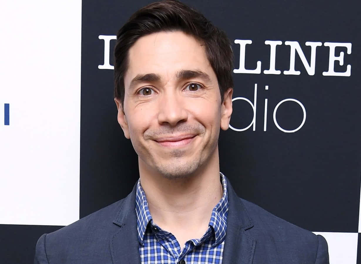 Justin Long 桌布 壁紙 — 一名深色頭髮、帶有含蓄微笑的男子，在品牌背景前穿著藍色西裝外套，內搭格仔恤衫。