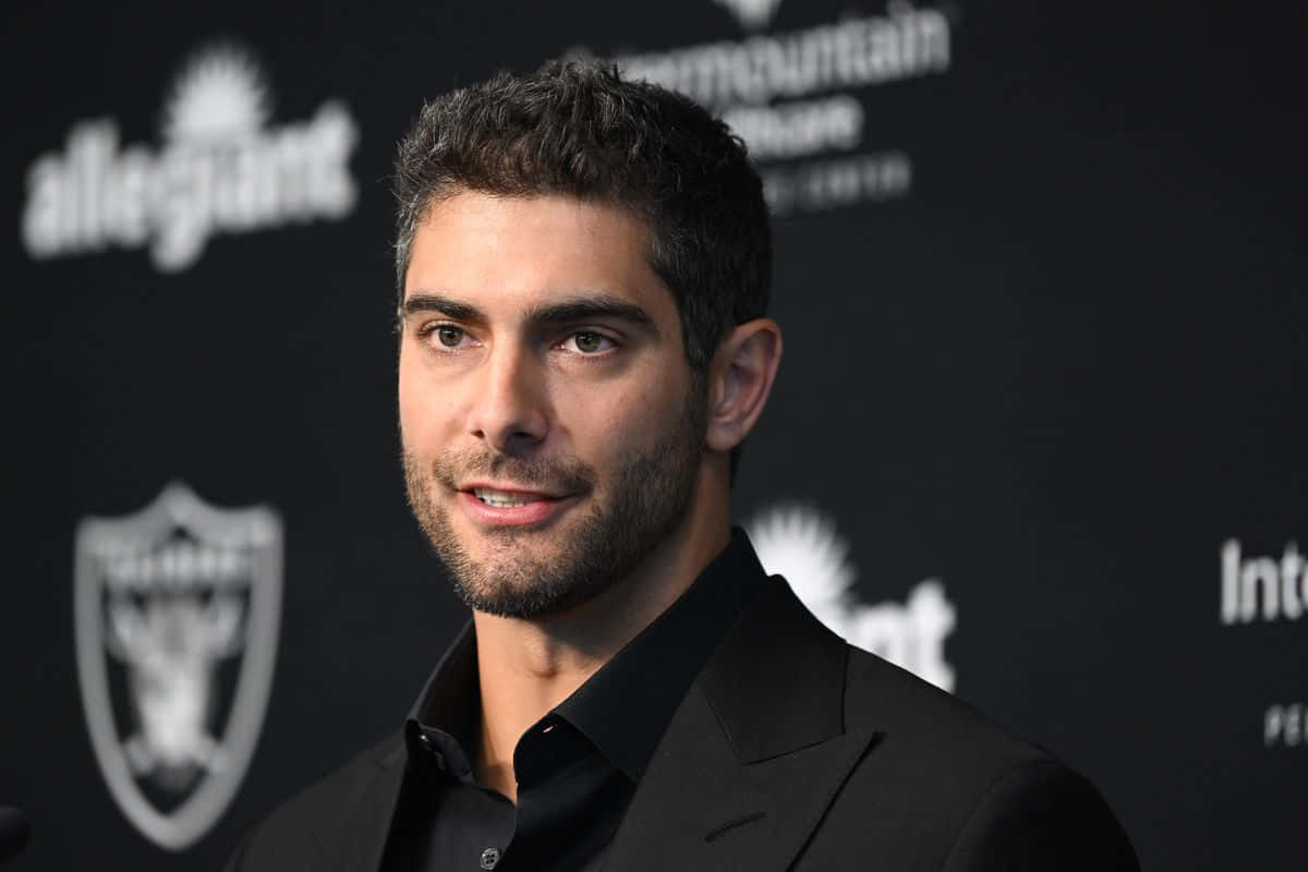 Jimmy Garoppolo 桌布 壁紙 — 一名深色頭髮和鬍鬚的男子，在帶有標誌的深色背景下穿著黑色西裝。
