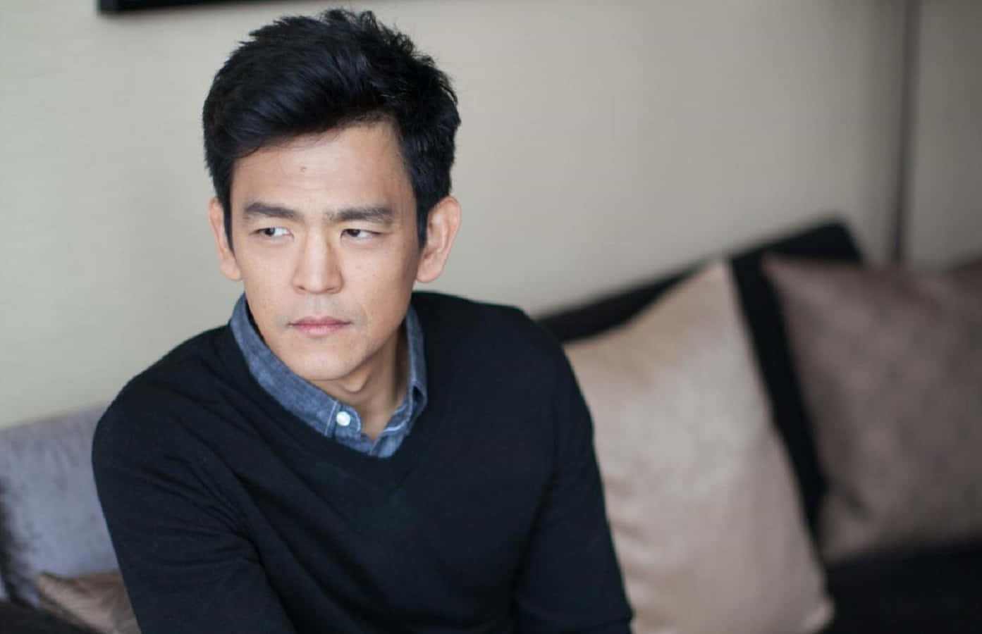 John Cho 桌布 壁紙 — 一名深色頭髮的男子坐在梳化上，身穿黑色毛衣內搭藍色恤衫，若有所思地望向一旁。