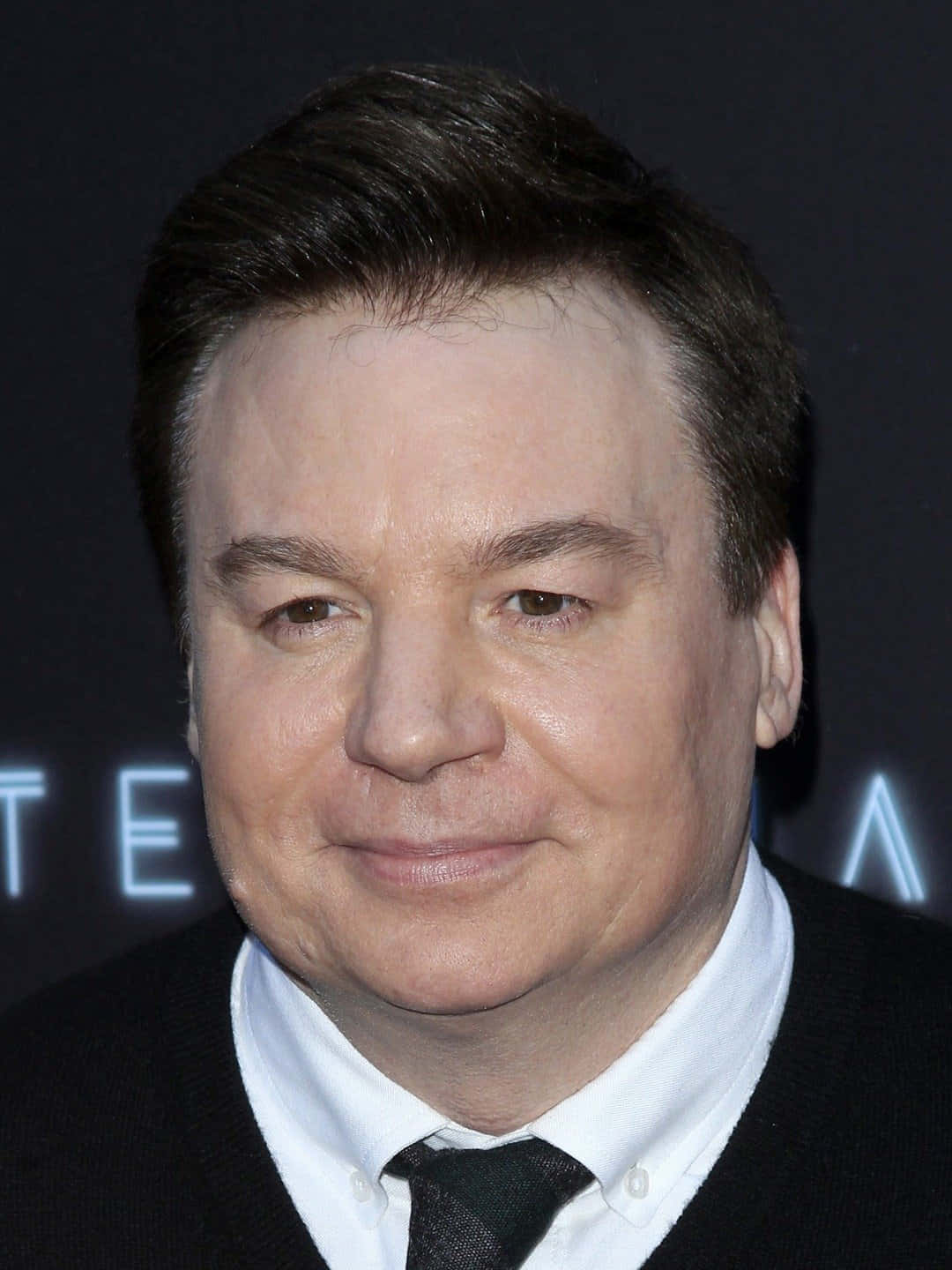 Mike Myers 桌布 壁紙 — 一名深色頭髮的男子穿著黑色西裝、白色襯衫和黑色領帶，在黑暗背景中帶有發光藍色字母的特寫肖像。