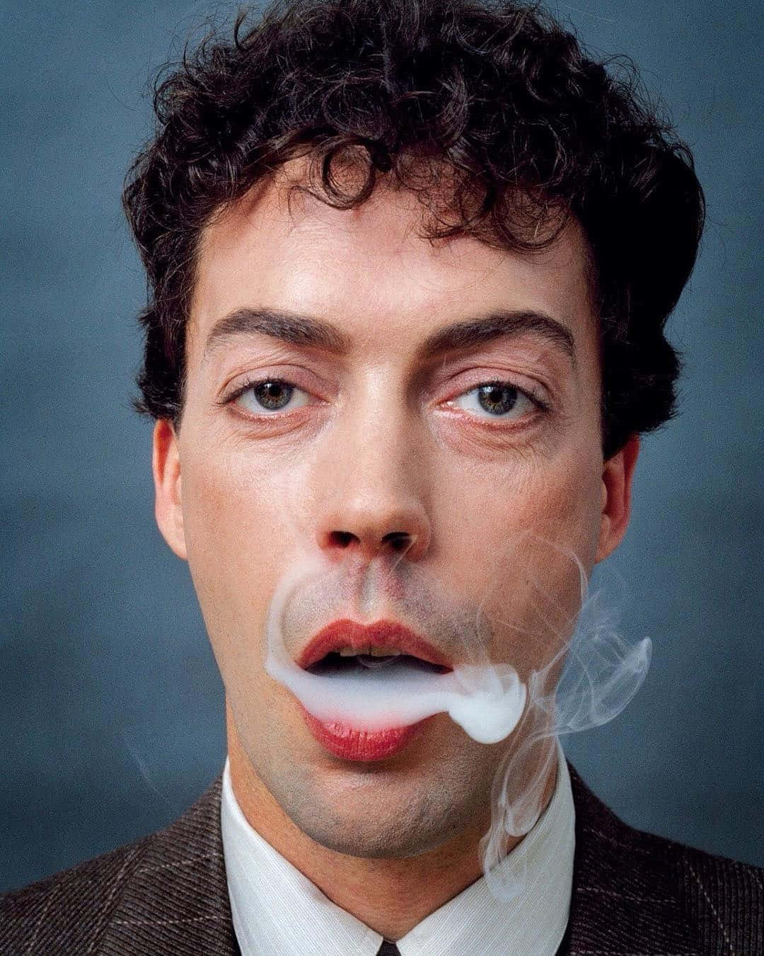 Tim Curry 桌布 壁紙 — 一名深色鬈髮、紅唇的男子在柔和的藍灰色攝影棚背景前呼出煙霧。