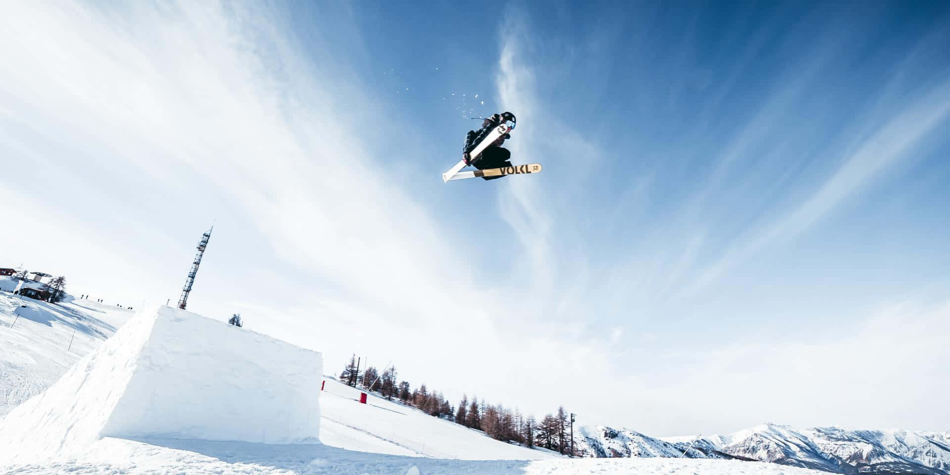 Skiing 桌布 壁紙 — 一名滑雪板運動員在晴朗的藍天中翱翔，下方是白雪覆蓋的山坡，遠處有山峰和稀疏的樹木。