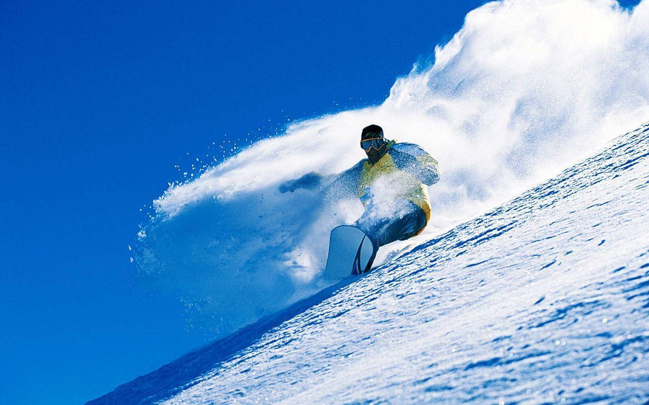 冬天 桌布 壁紙 — 一名滑雪板運動員在陡峭的雪坡上 carve 而下，將大量粉雪噴向清澈的藍天。