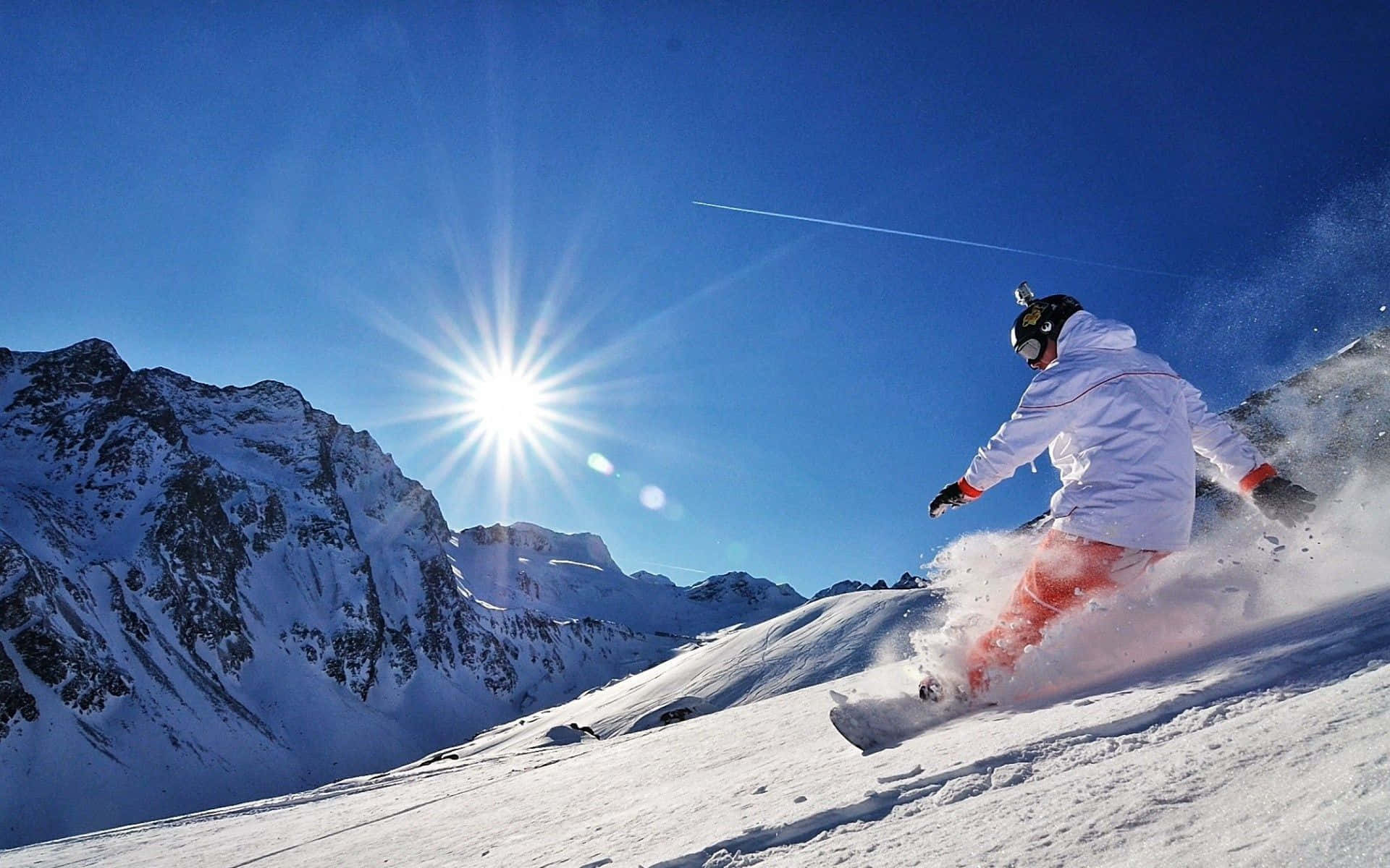 Winter Sports 桌布 壁紙 — 一名滑雪板運動員在陽光明媚的山坡上，穿過新鮮的粉雪，背景是白雪覆蓋的山峰和湛藍的天空。