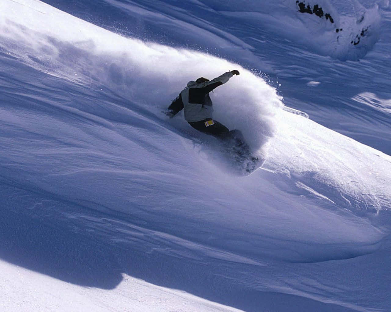 Winter Sports 桌布 壁紙 — 一名滑雪板運動員在陽光明媚的山坡上穿過深粉雪，將白色的雪花戲劇性地噴灑到空中。
