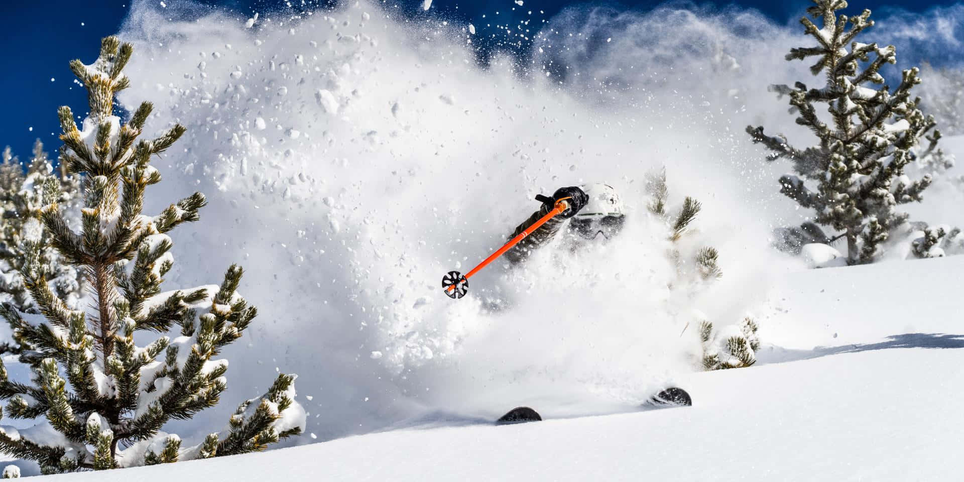 Skiing 桌布 壁紙 — 一名滑雪者在新鮮的粉雪中滑行，揚起大量雪花，映襯著清澈的藍天。