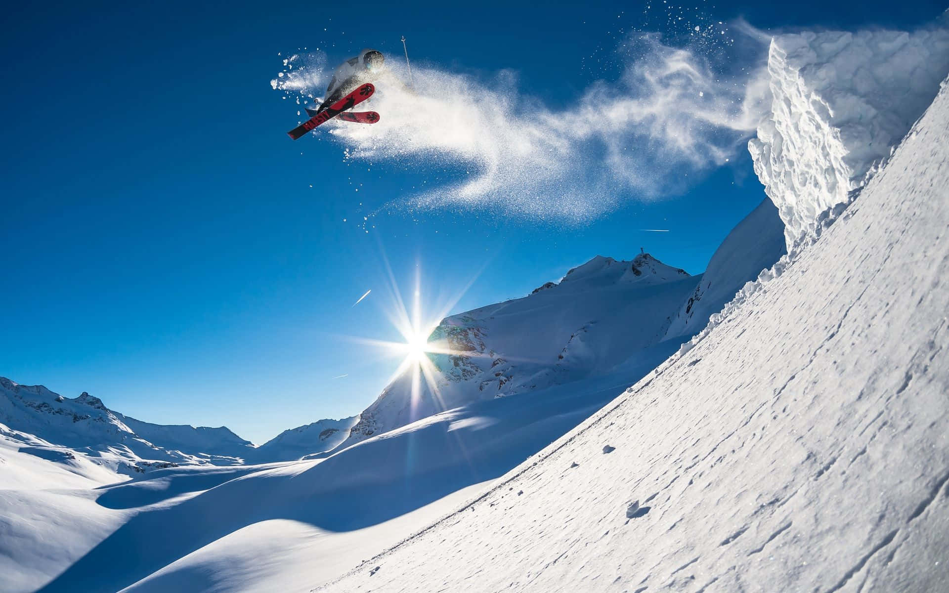 Skiing 桌布 壁紙 — 一名滑雪者在晴朗的藍天中，飛越白雪覆蓋的山坡，陽光耀眼。