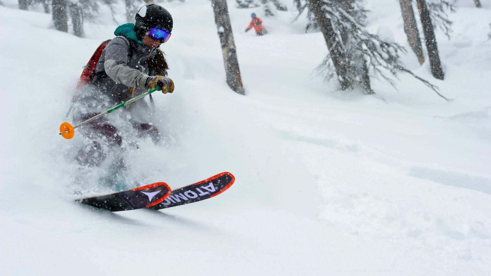 Skiing 桌布 壁紙 — 一名滑雪者在白雪覆蓋的森林中穿過深粉雪，背景可見樹木和另一名滑雪者。