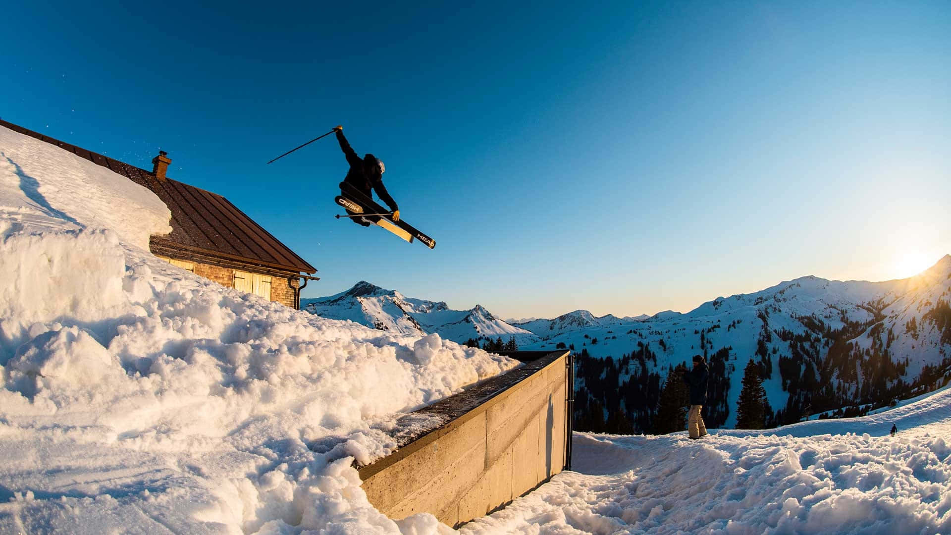 Skiing 桌布 壁紙 — 一名滑雪者在金色日落時分，於白雪覆蓋的山間小屋上方，在清爽的冬日空氣中翱翔。