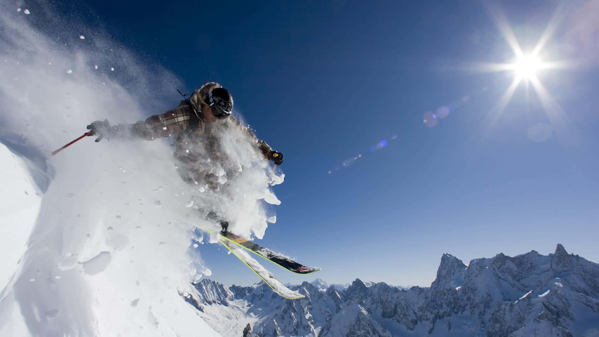 Skiing 桌布 壁紙 — 一名滑雪者在陽光明媚的山坡上劃過新雪，雪花飛濺到清澈的藍天中。