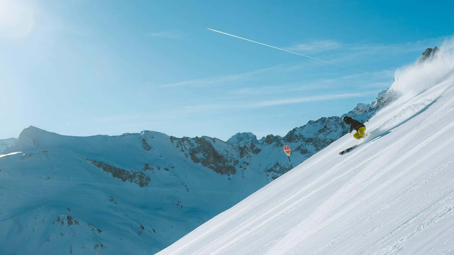 Skiing 桌布 壁紙 — 一名滑雪者在陽光明媚的雪坡上，在鋸齒狀的山峰和清澈的藍天下，穿過新鮮的粉雪。