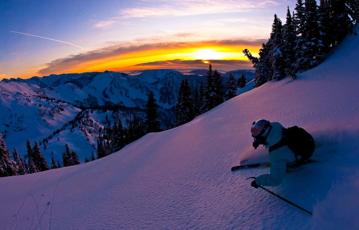 Winter Sports 桌布 壁紙 — 一名滑雪者沿著白雪覆蓋的山坡滑下，夕陽在遠峰後落下，將溫暖的橙色光線灑在雪地上。