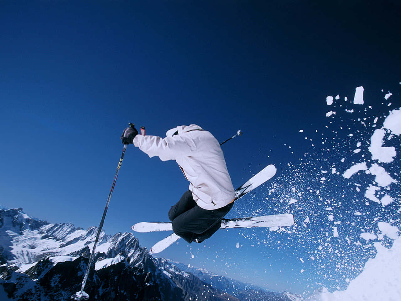 Skiing 桌布 壁紙 — 一名滑雪運動員在清澈的藍天中翱翔，越過白雪覆蓋的山峰，滑雪板濺起雪花。
