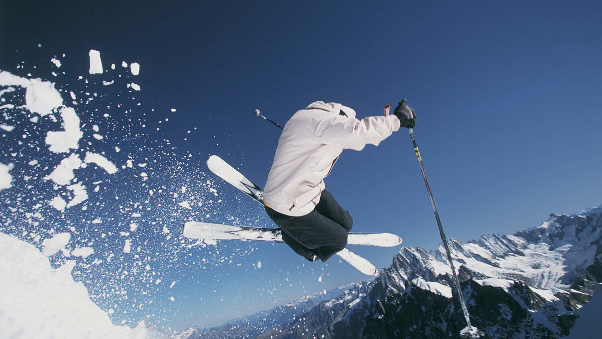 Skiing 桌布 壁紙 — 一名滑雪運動員在白雪覆蓋的山峰上空，劃過清澈的藍天，滑雪板濺起雪花。