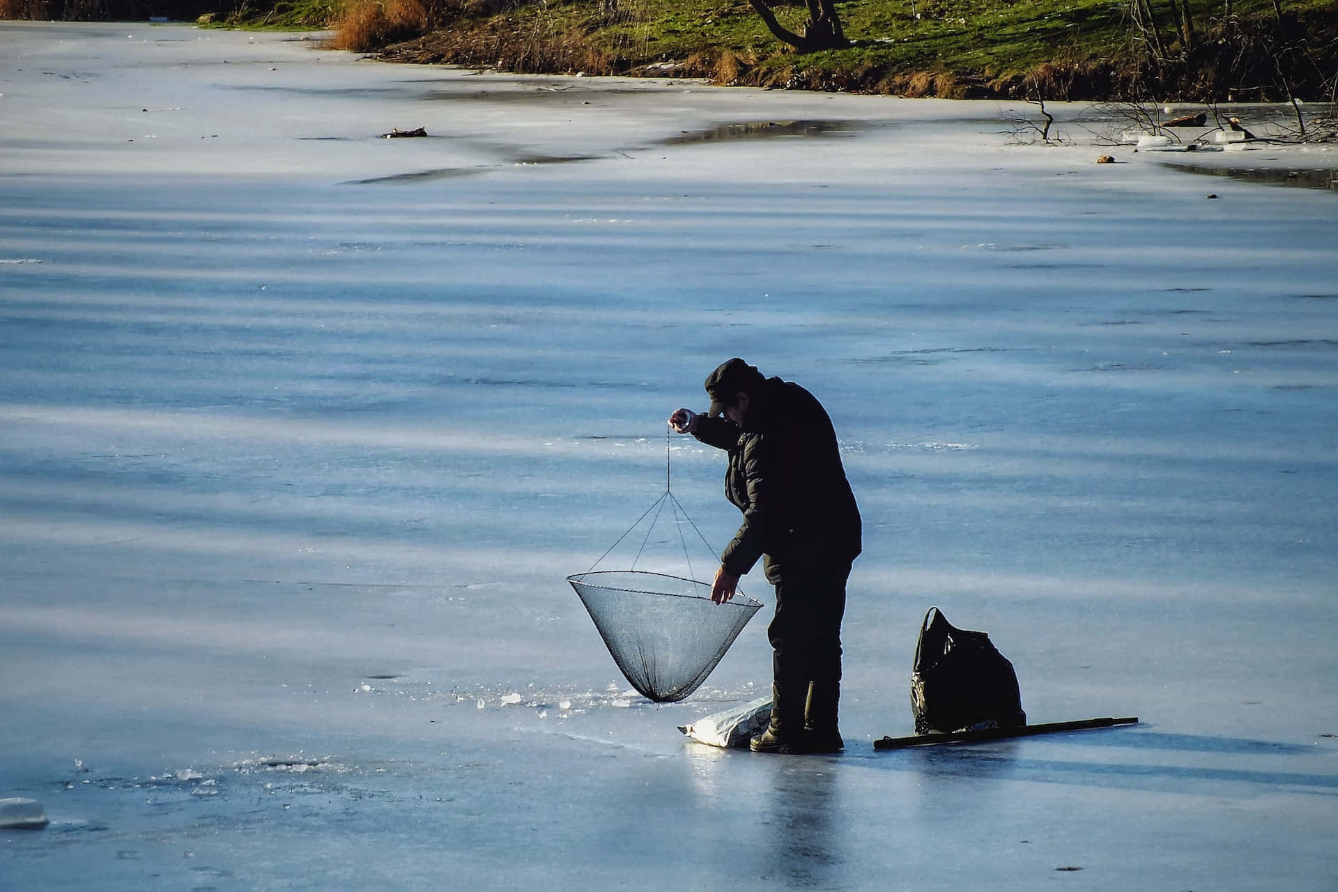 Ice Fishing 桌布 壁紙 — 一名漁夫站在結冰的湖面上，在明亮的陽光下，手持漁網舉在冰面上方，附近有一個背包。