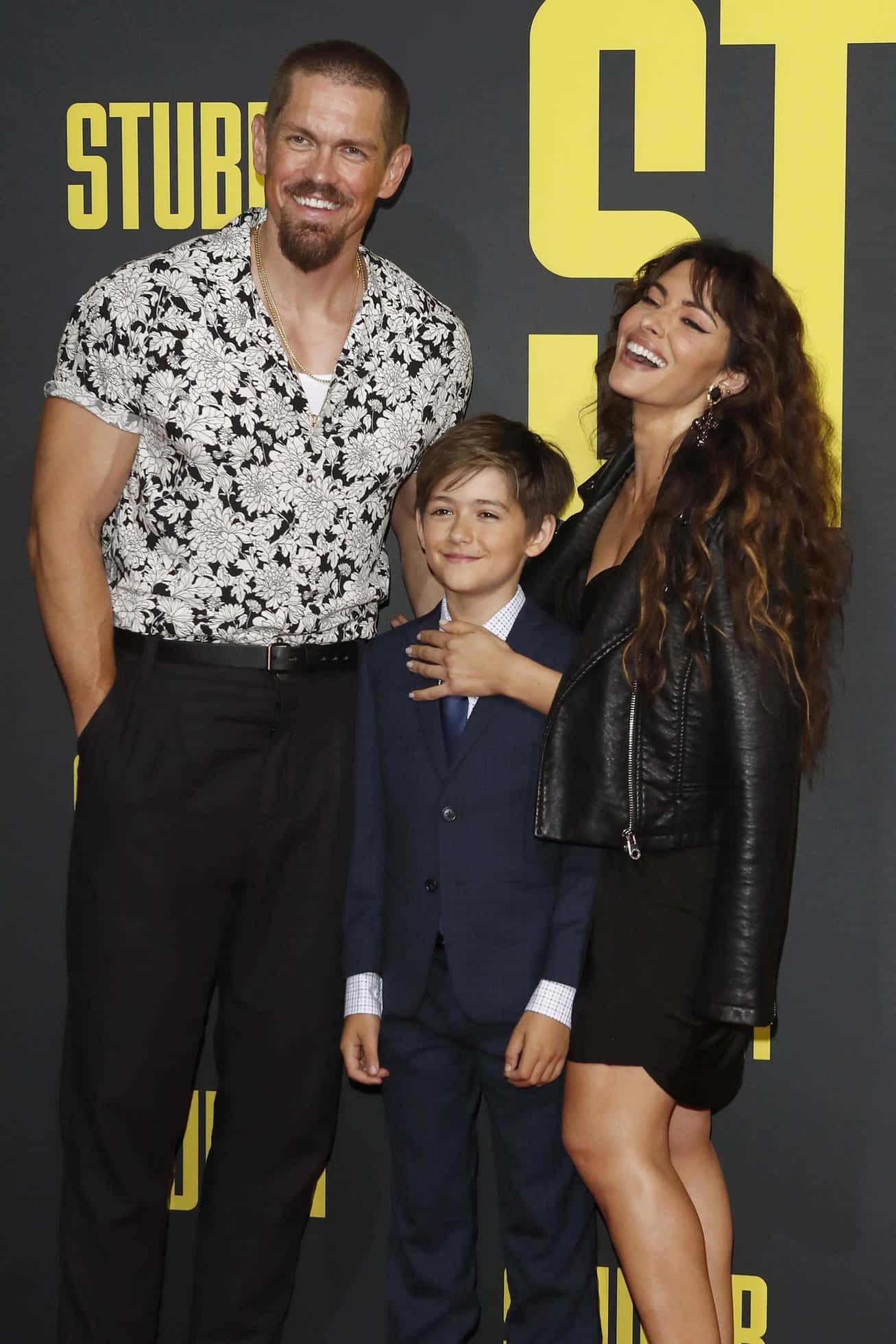 sarah shahi 桌布 壁紙 — 一名男子、女子和小孩在電影首映禮上，在搶眼的黃色背景前一起微笑。